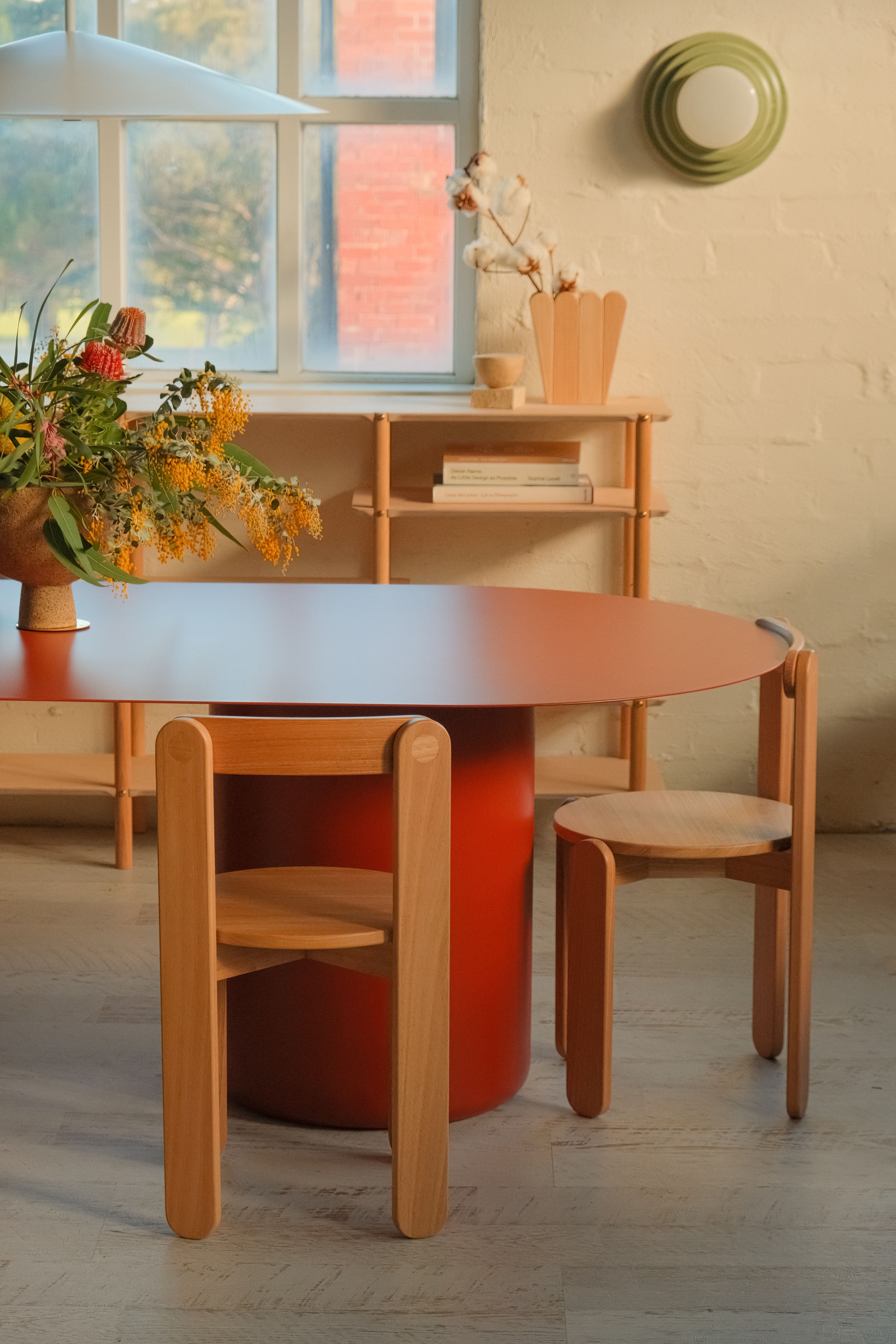A red oval dining table in a sunny room with timber chairs and native flowers. 