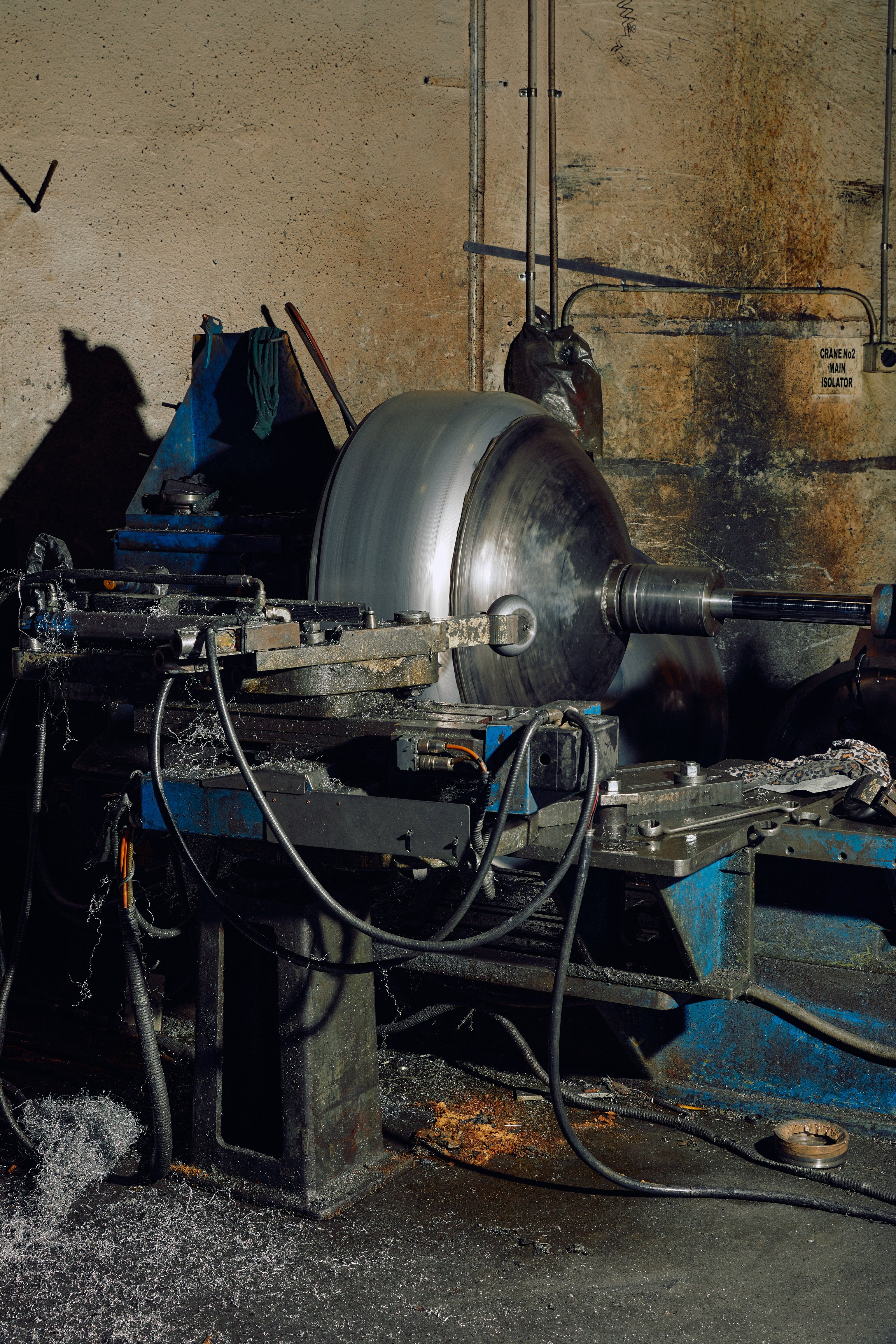 A large metal shape being spun on a lathe by a roller in a messy industrial factory