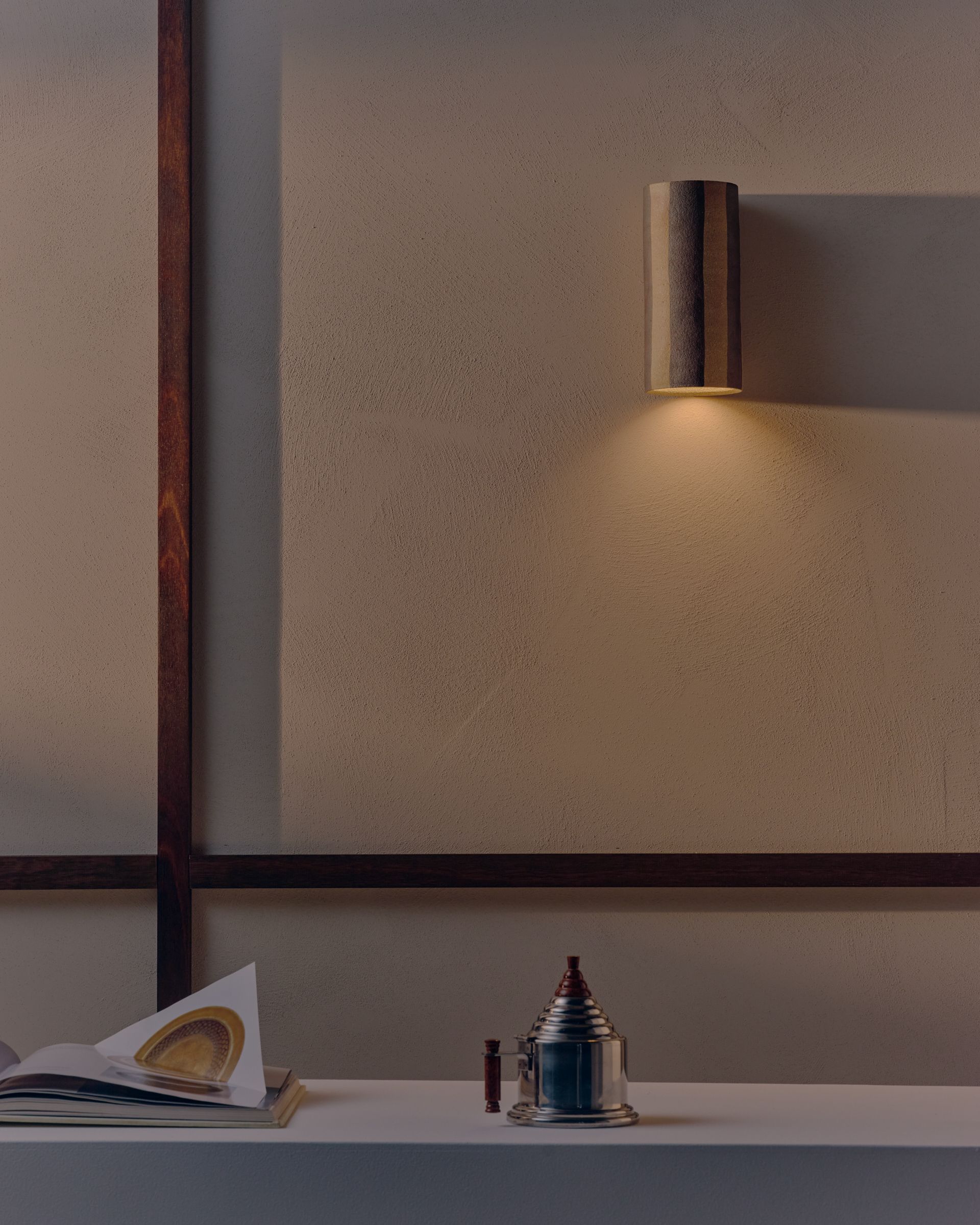 A striped cylindrical ceramic wall light throws a warm glow over the textured cream wall. Below is an antique coffee pot and an open book. 