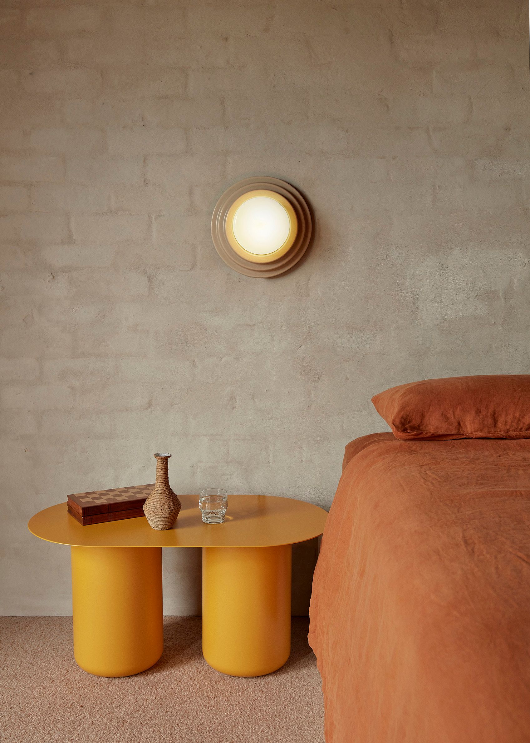 A glowing Honey Wall Light mounted to a rendered brick wall beside a bed and a yellow Sequence 02 coffee table