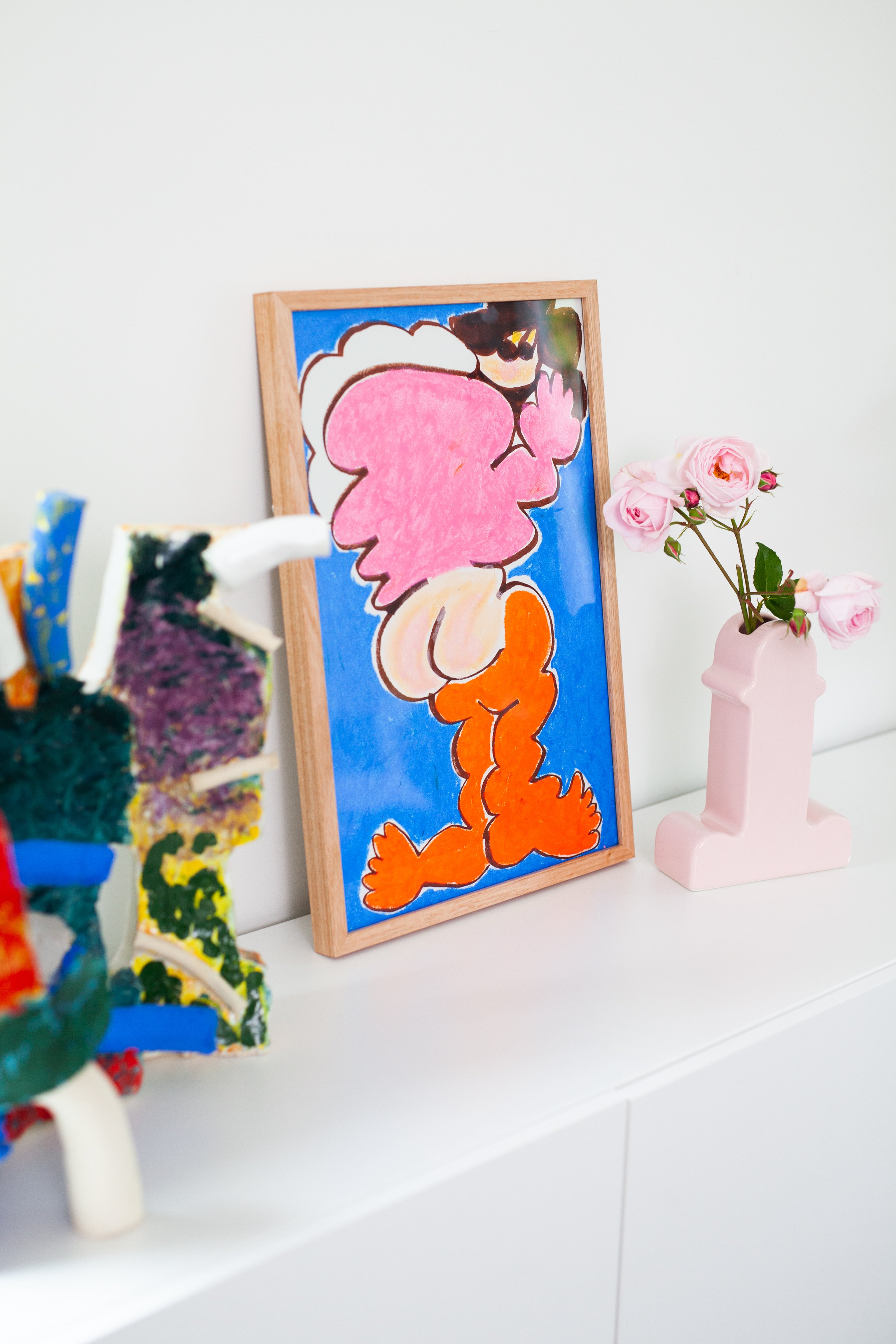 A colourful painting sitting on a white cabinet leaning against a white wall. Also on the cabinet are some vases of flowers