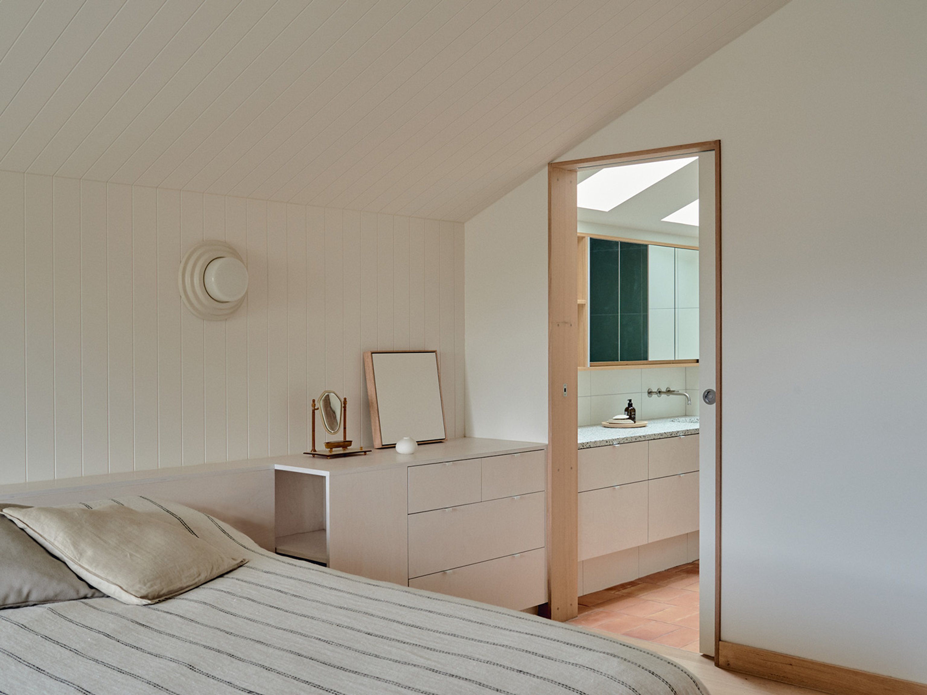 A bedroom with white walls and a white ceramic wall light attached to the back wall. The bedroom is looking into an ensuite