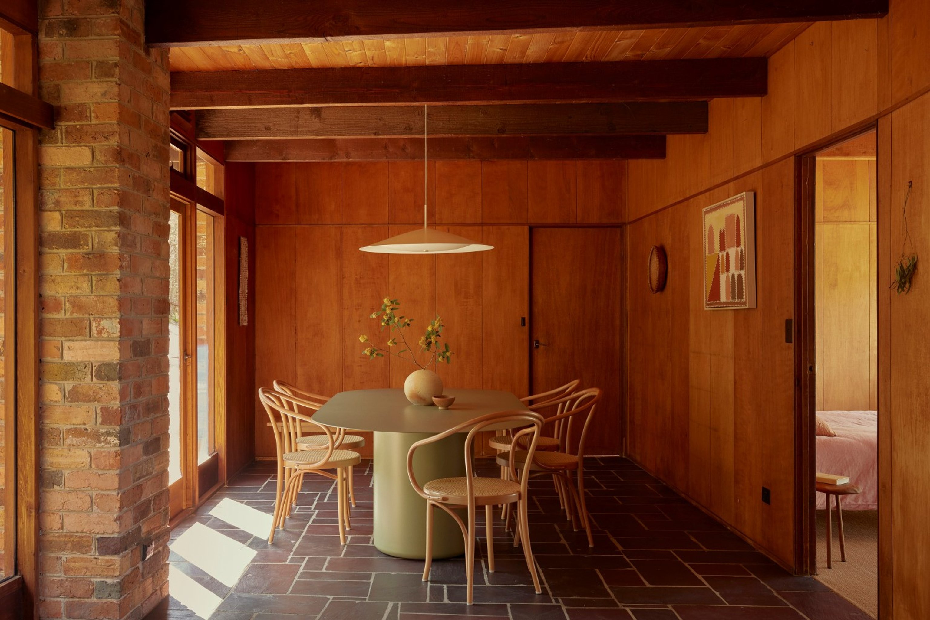 A green oval dining table in a brick and timber clad room with slate floors and timber ceilings. A round Broad pendant hangs over the dining table and light is pouring in through the windows.