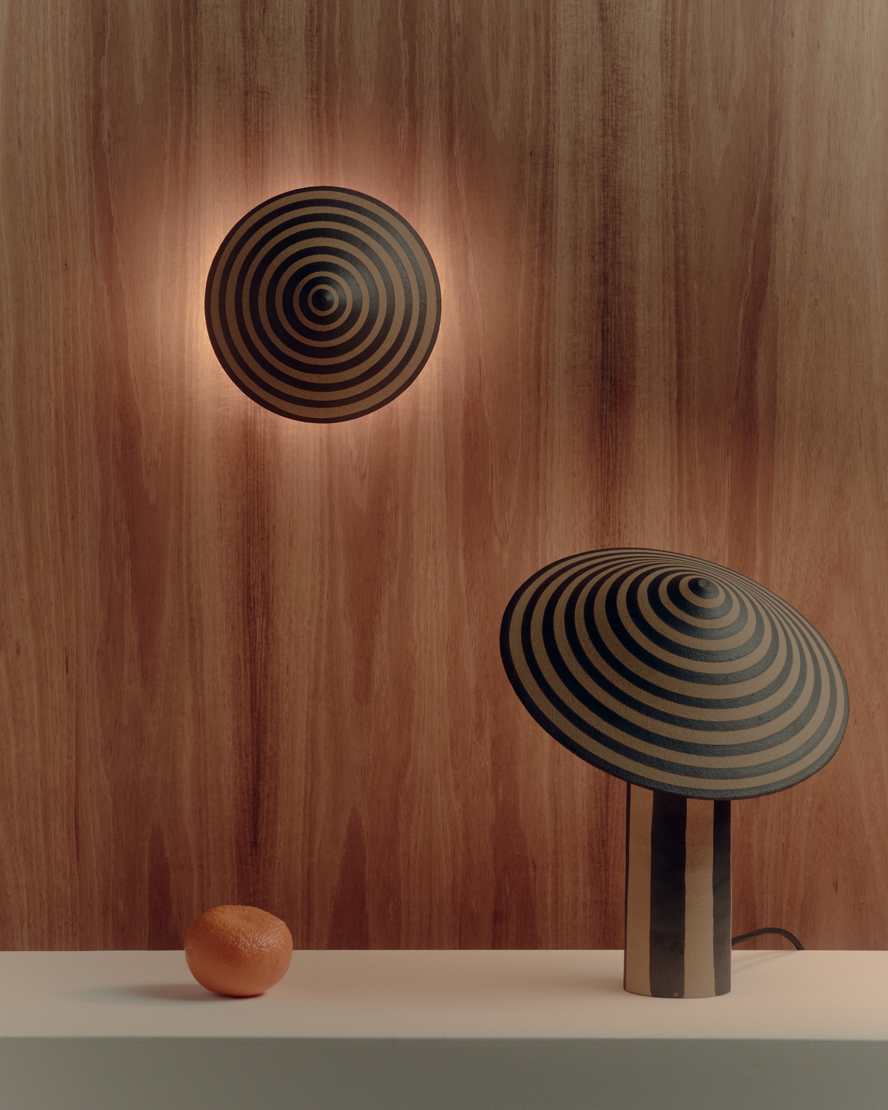 A striped ceramic table lamp with a wide striped base and hand-painted circular shade sitting next to an orange against a timber wall with a striped ceramic wall light