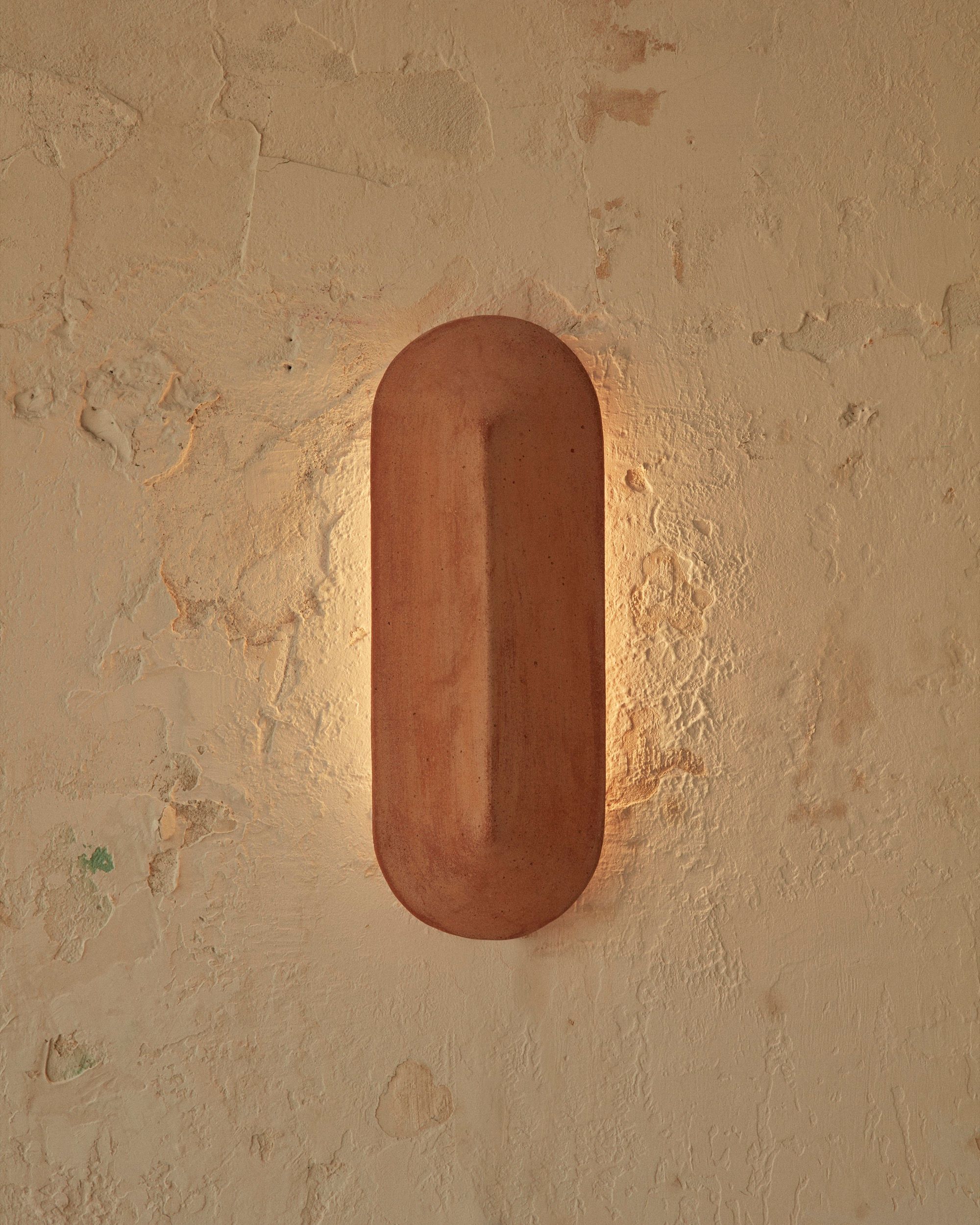 A Pinch 400 elongated wall light made of terracotta, against a white brick background. Light is illuminating the wall it is fixed to