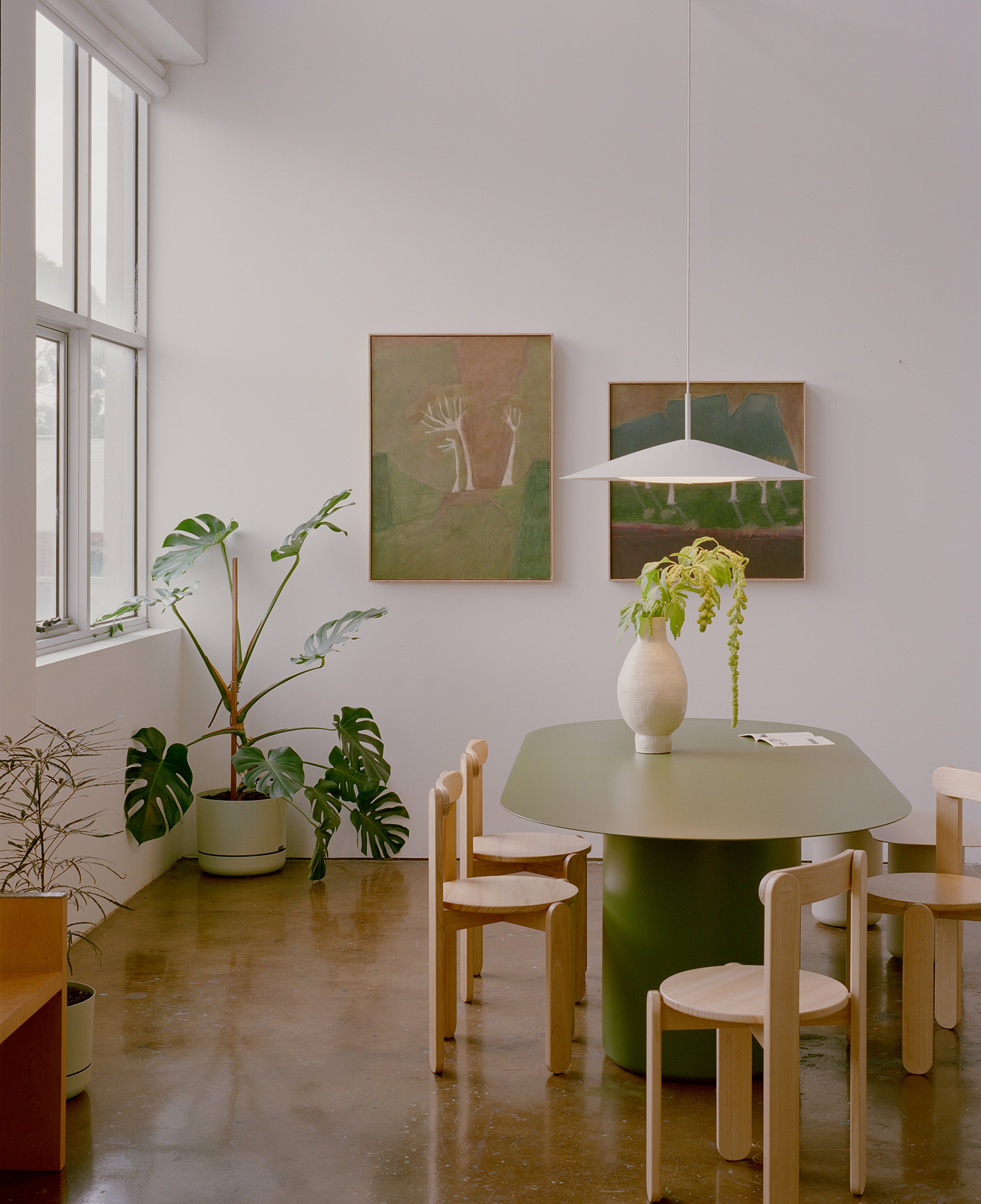 A green oval table with a broad white pendant light sitting above. The table has a large vase with greenery and there are timber chairs surrounding the table. Muted artworks hang on the white wall behind and there is a window and indoor plant.