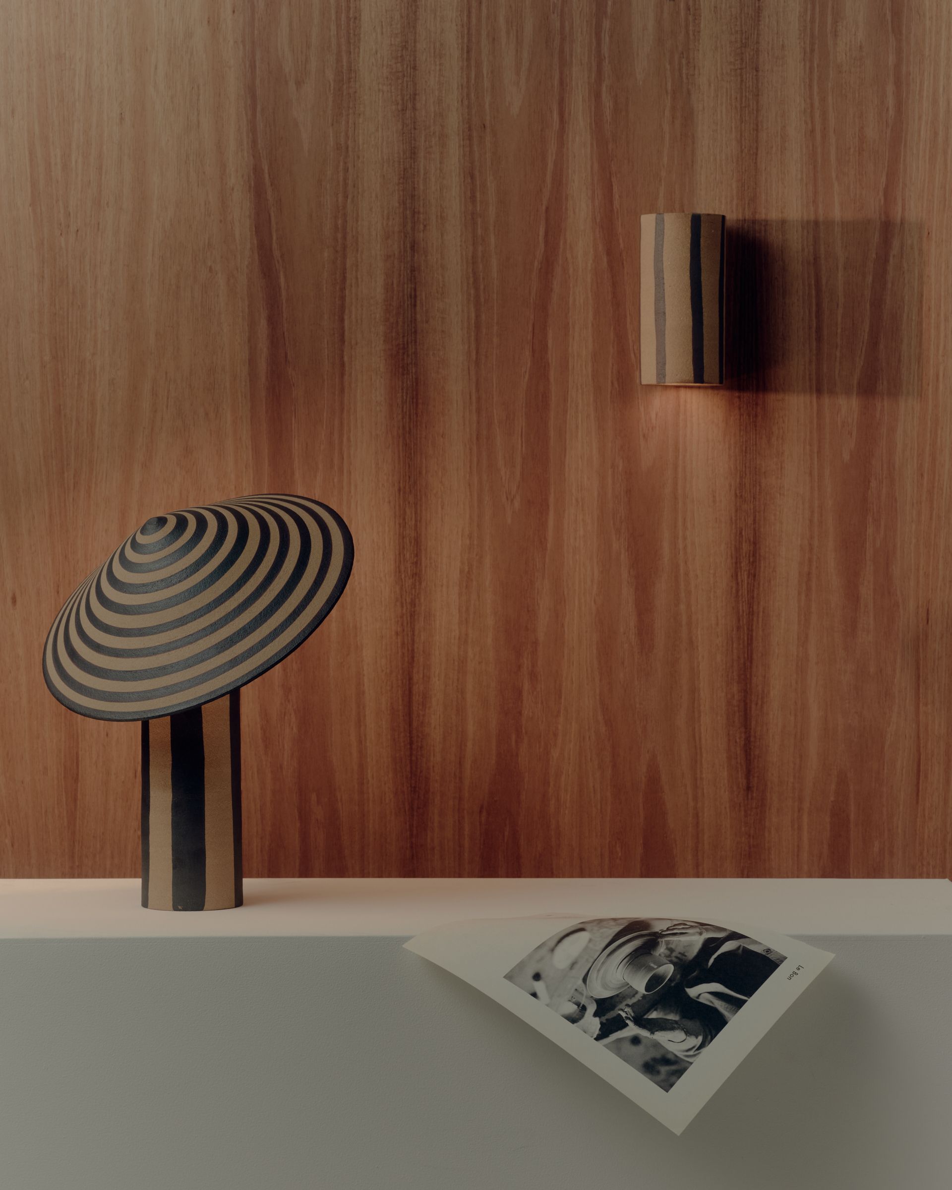 A large striped ceramic table lamp sitting on a fabric plinth with a ceramic striped wall lamp attached to a dark timber wall