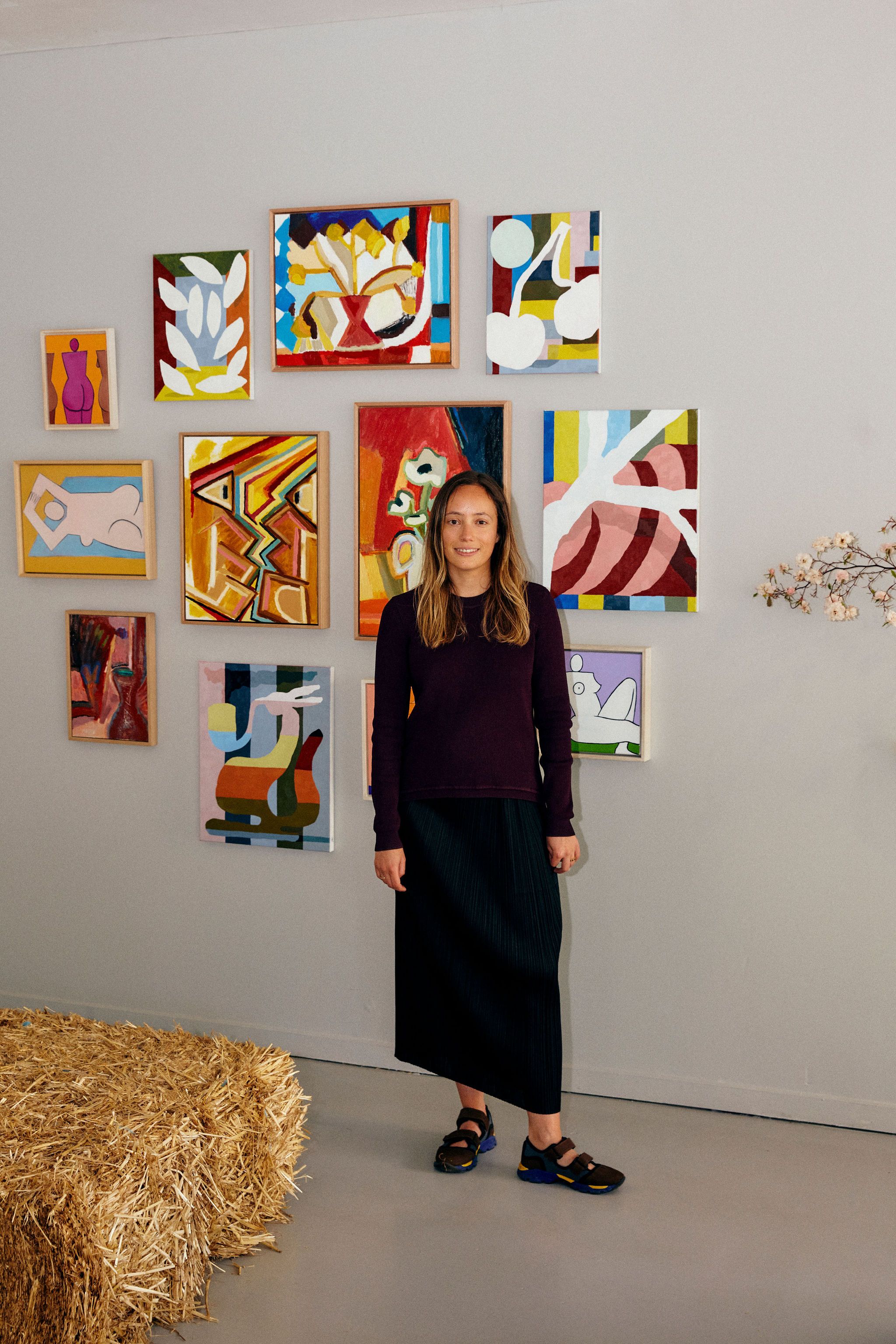 A portrait of Lily Mora standing in front of a group of paintings on a white wall