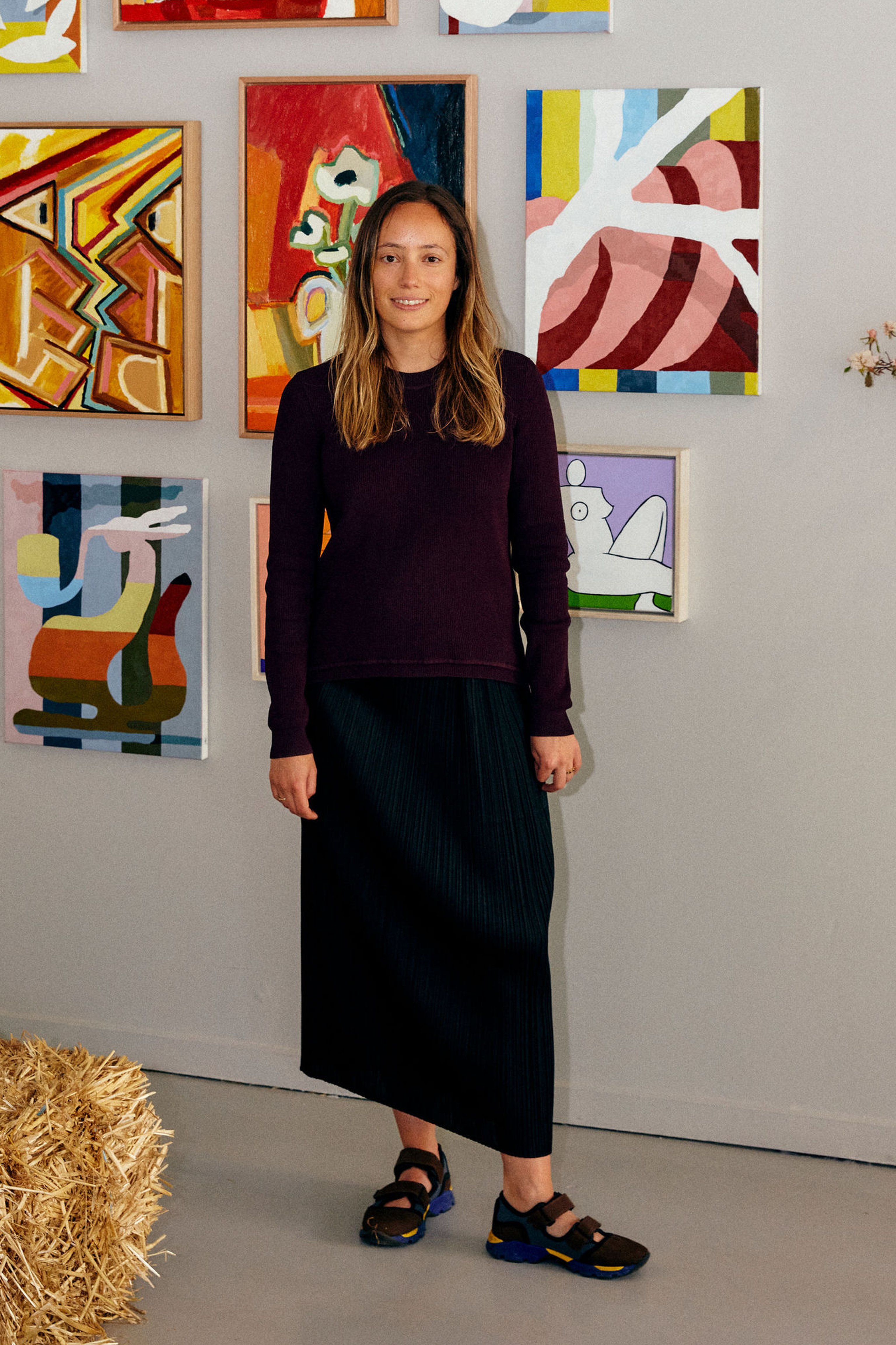 A portrait of Lily Mora standing in front of a group of paintings on a white wall