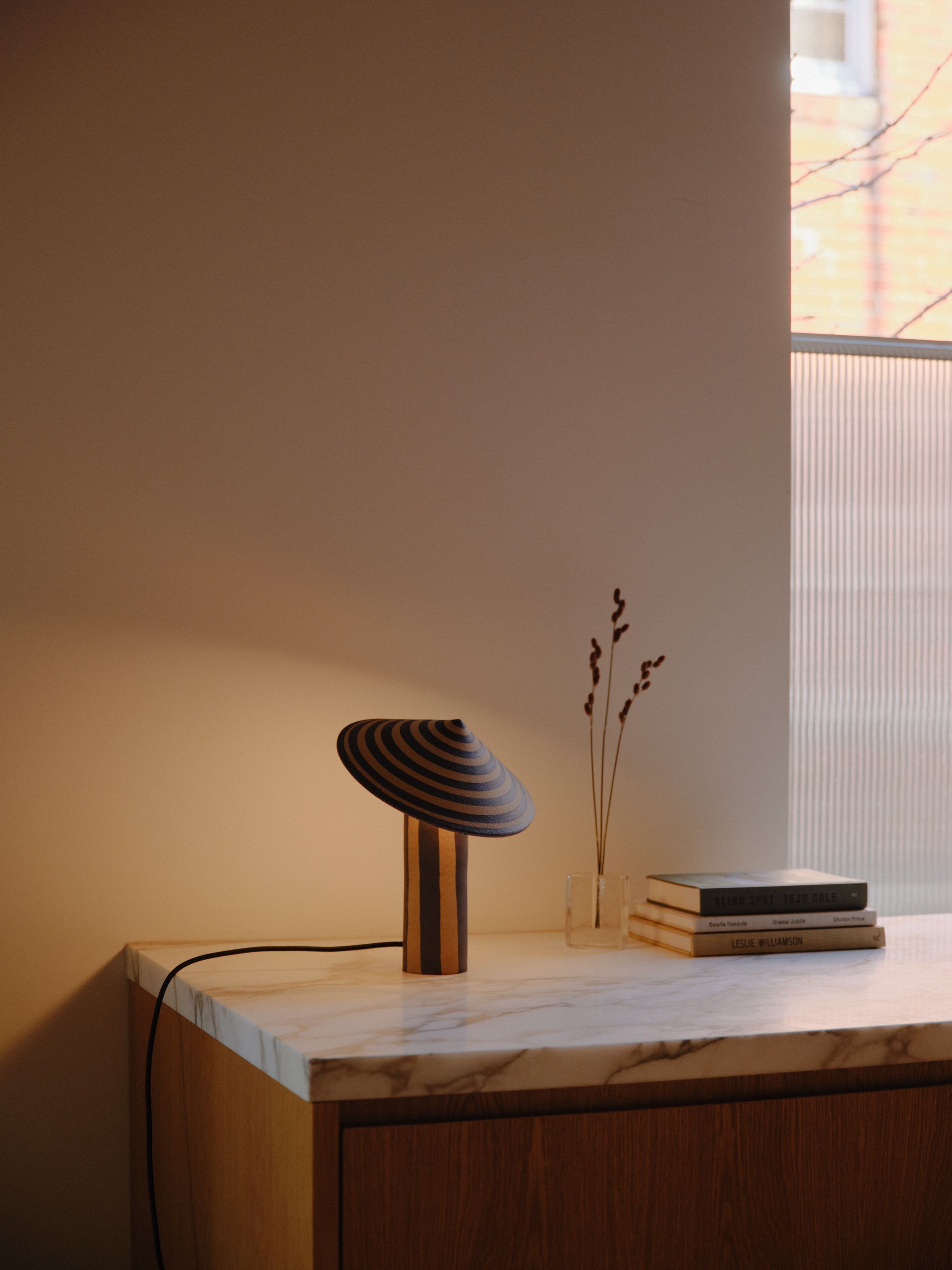 A striped ceramic Dancer Table Lamp sits on a marble kitchen bench next to some flowers and books with a window letting in some soft natural light