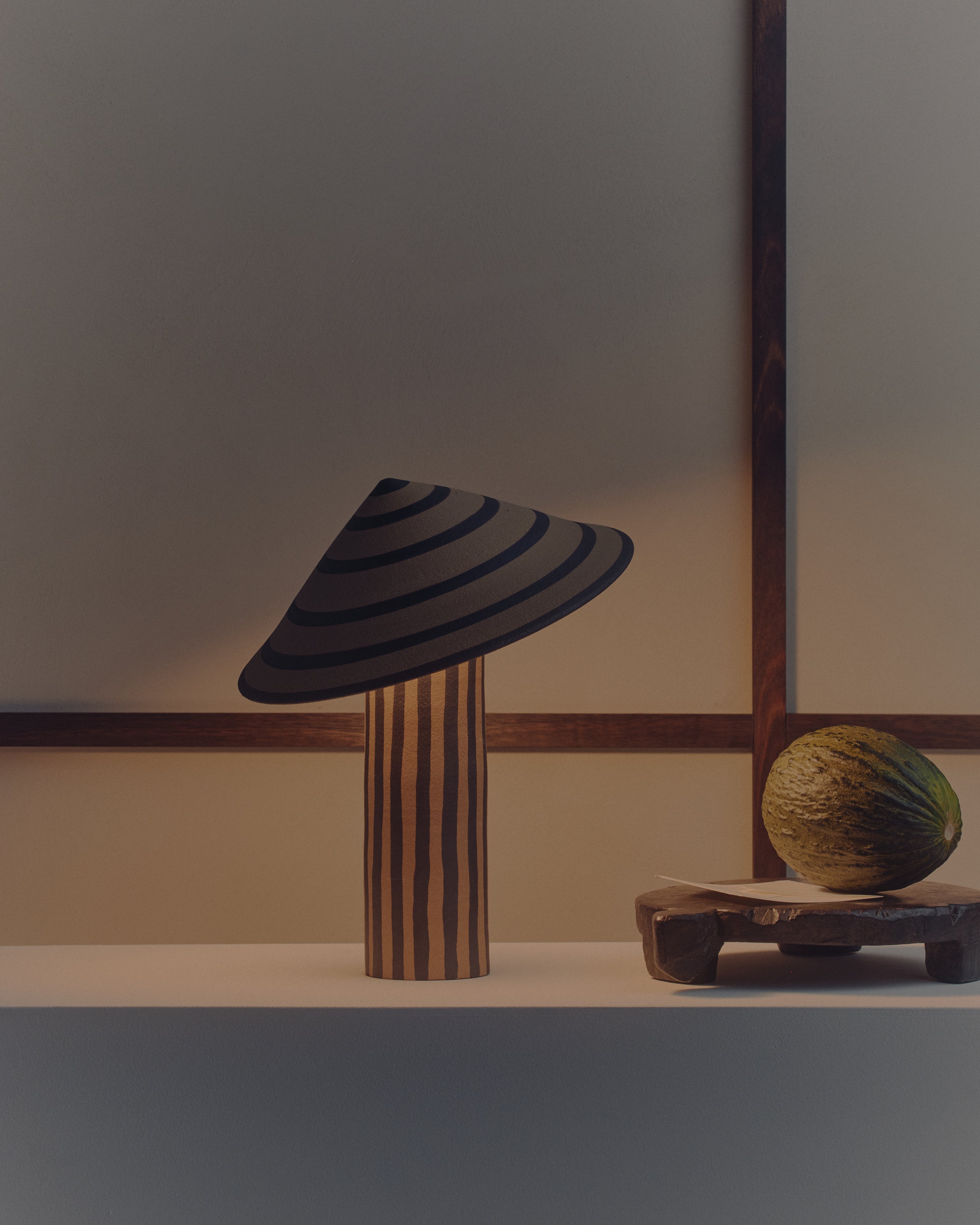 A striped ceramic table lamp with the shade on an angle sitting in front of a white wall with a melon next to it.