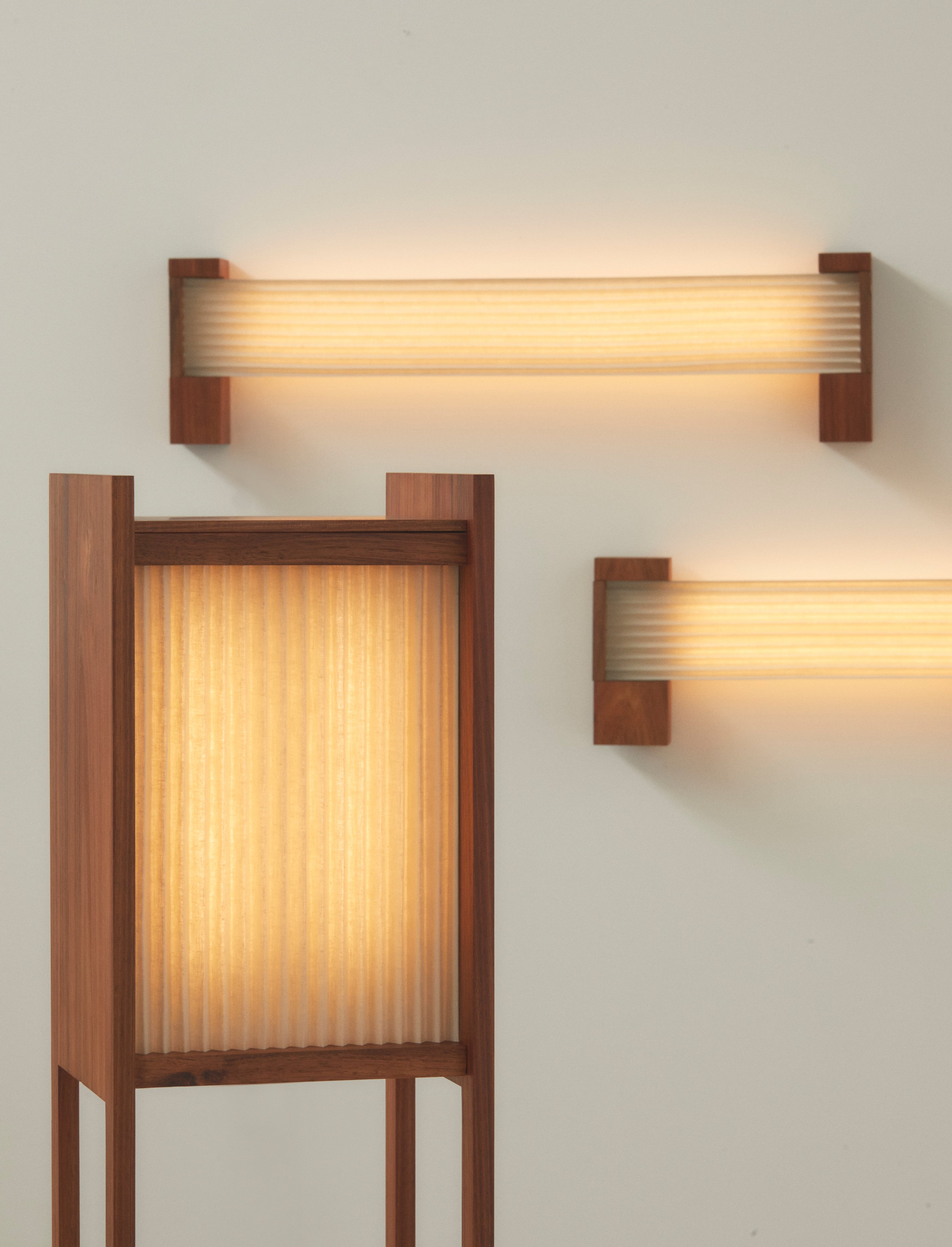 Timber and pleated linen floor lamp and wall lights on a white background. They have a warm glow.