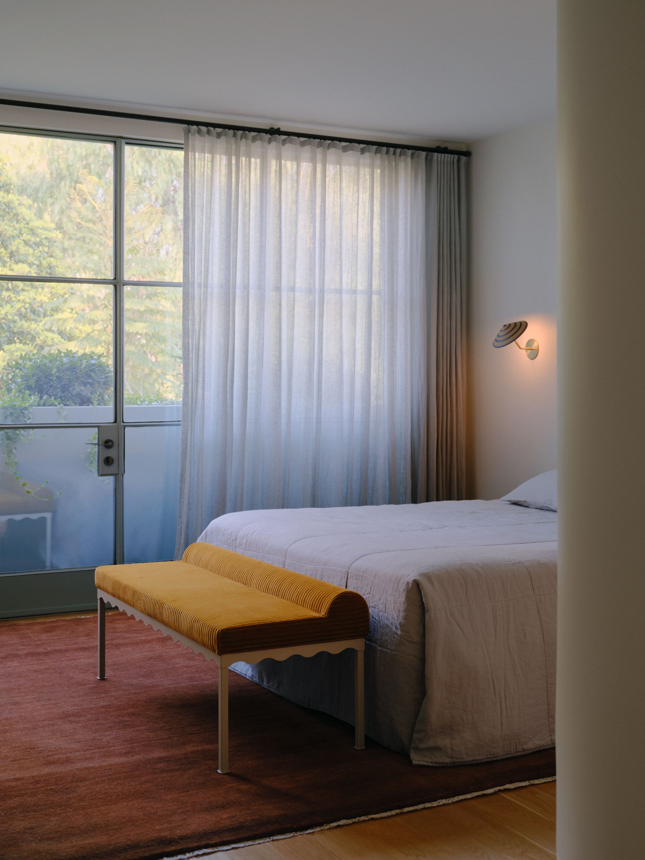 A bedroom setting with a ceramic striped Dancer Angled Wall Light and a Bellini Upholstered Bench at the end of the bed. Windows look out onto greenery through sheer curtains. 