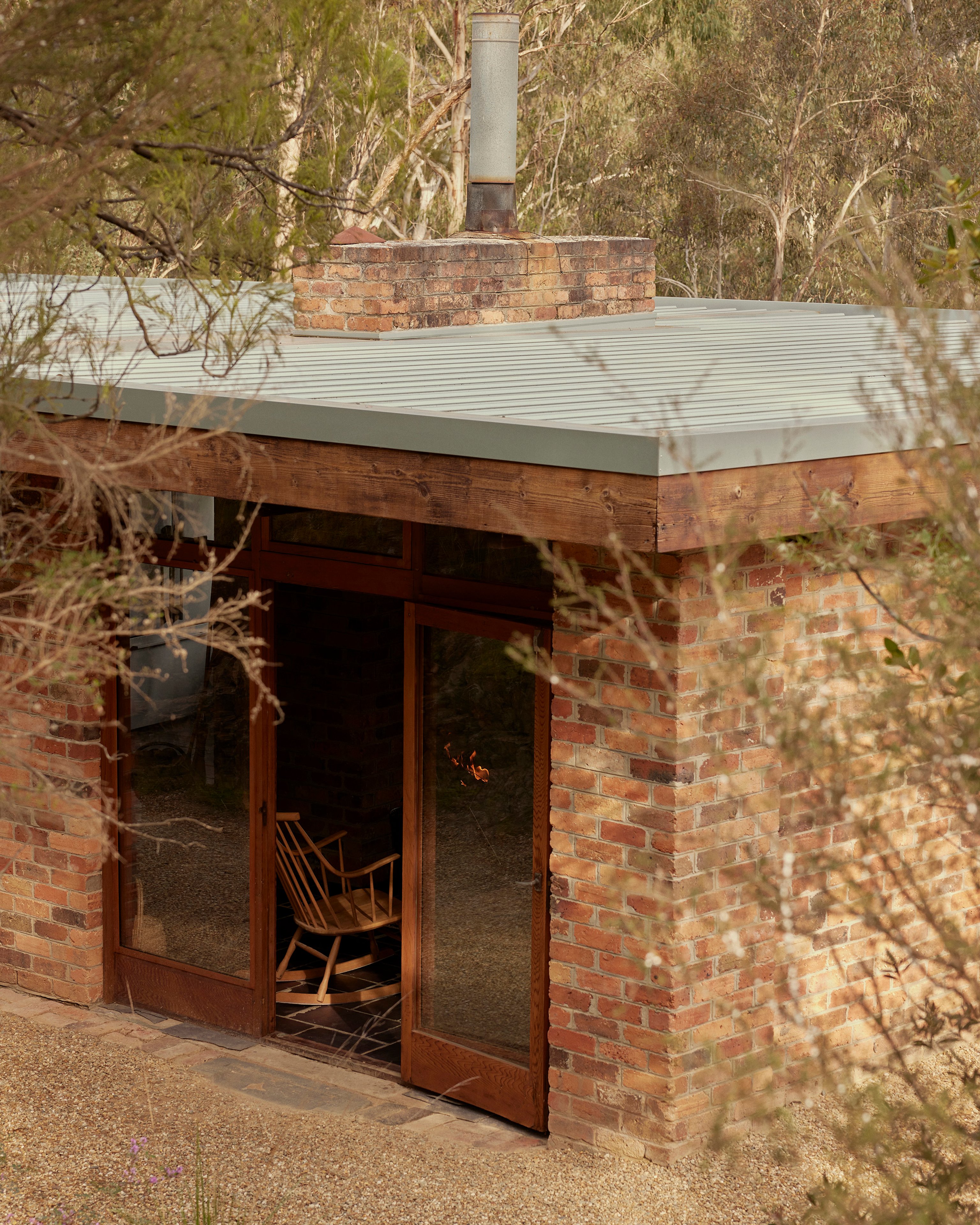 A modernist brick building with timber doorframes sits amongst trees. There is a rocking chair inside on the slate floor.