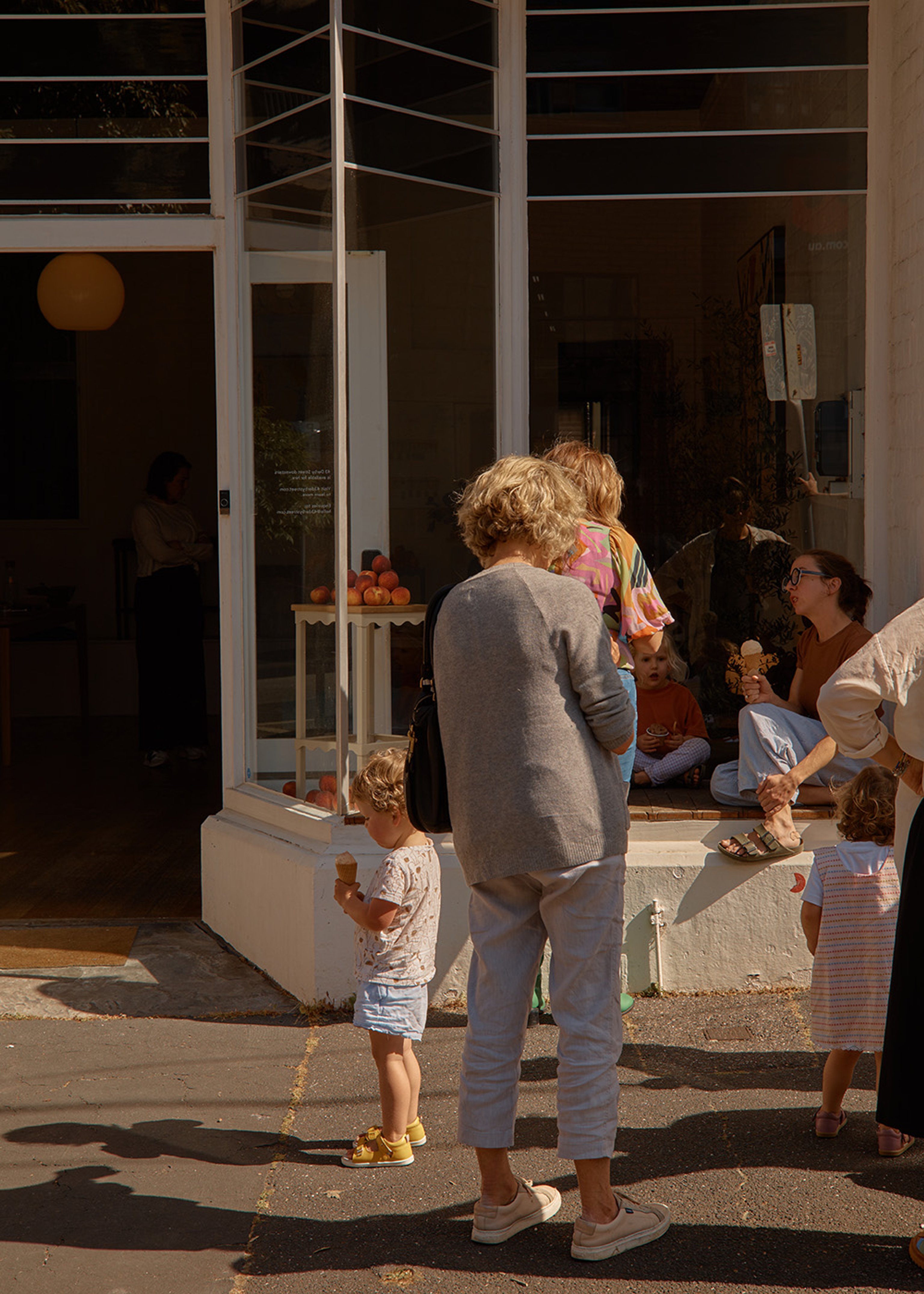 The outside of a small shop. The shop has a open door and large glass windows full of people. There is a mother and two children in the centre of the image