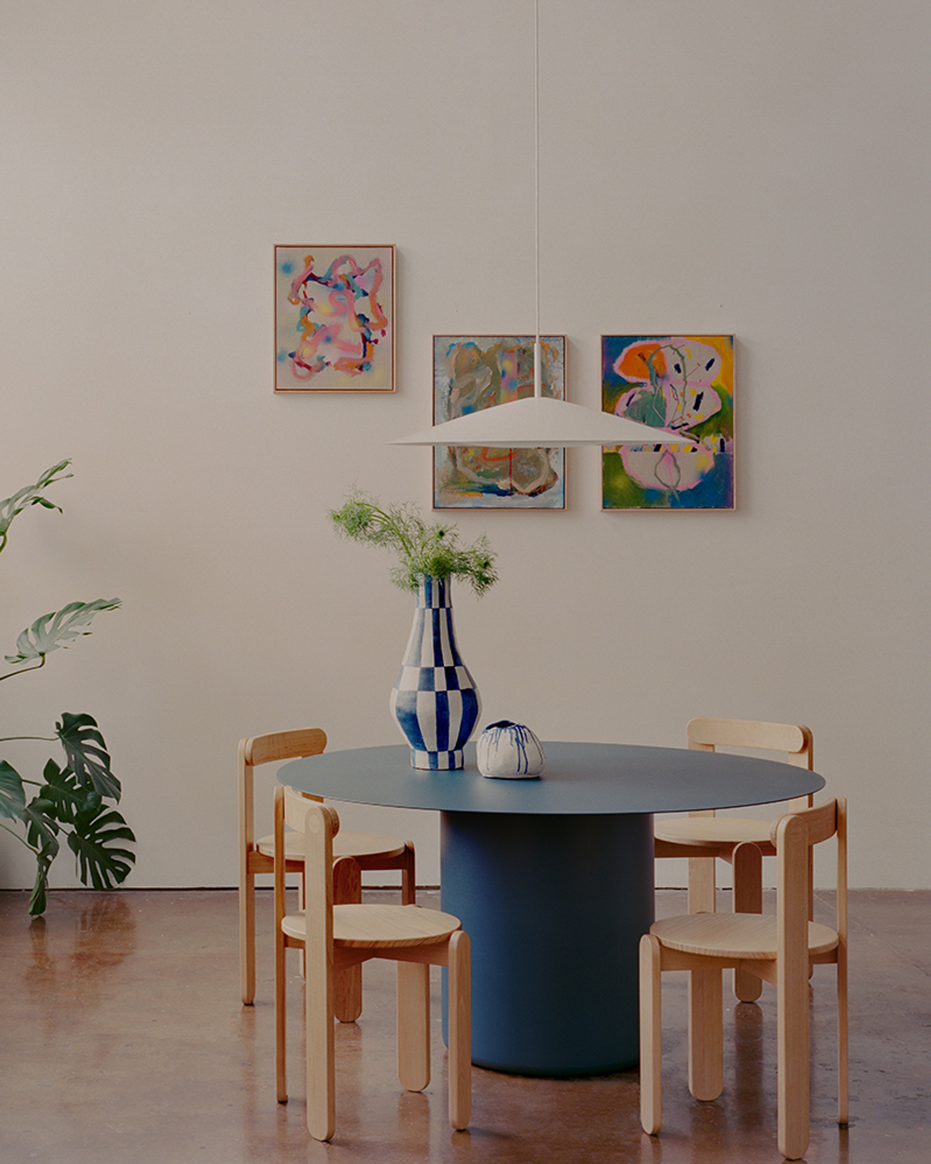 A round Sequence Dining Table in a Deep Ocean colour surrounded by timber chairs. A white Broad Pendant Light hangs above and there is art hanging on the wall behind.