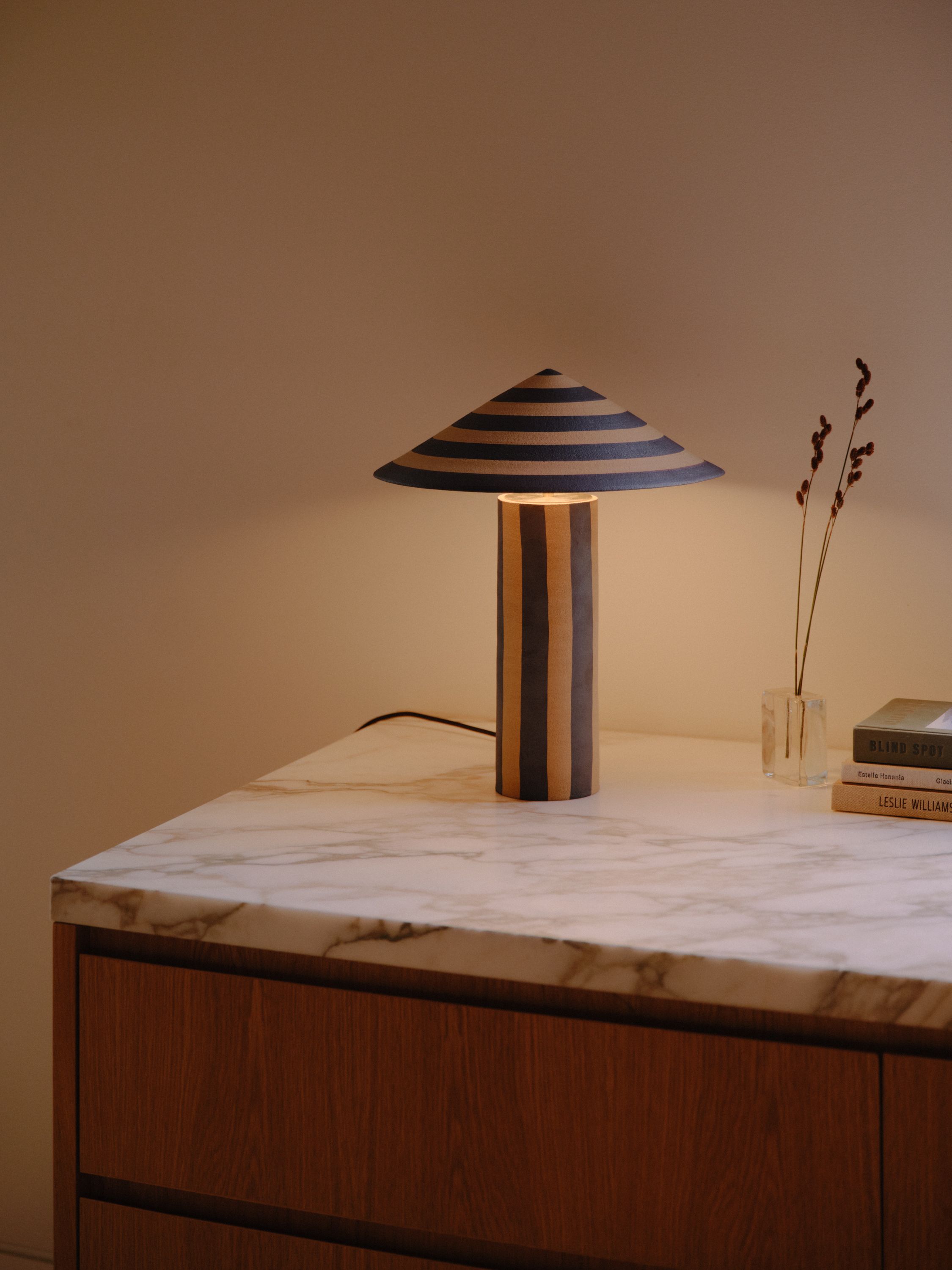 A striped ceramic Dancer Table Lamp sits on a marble kitchen bench next to some flowers and books with a window letting in some soft natural light