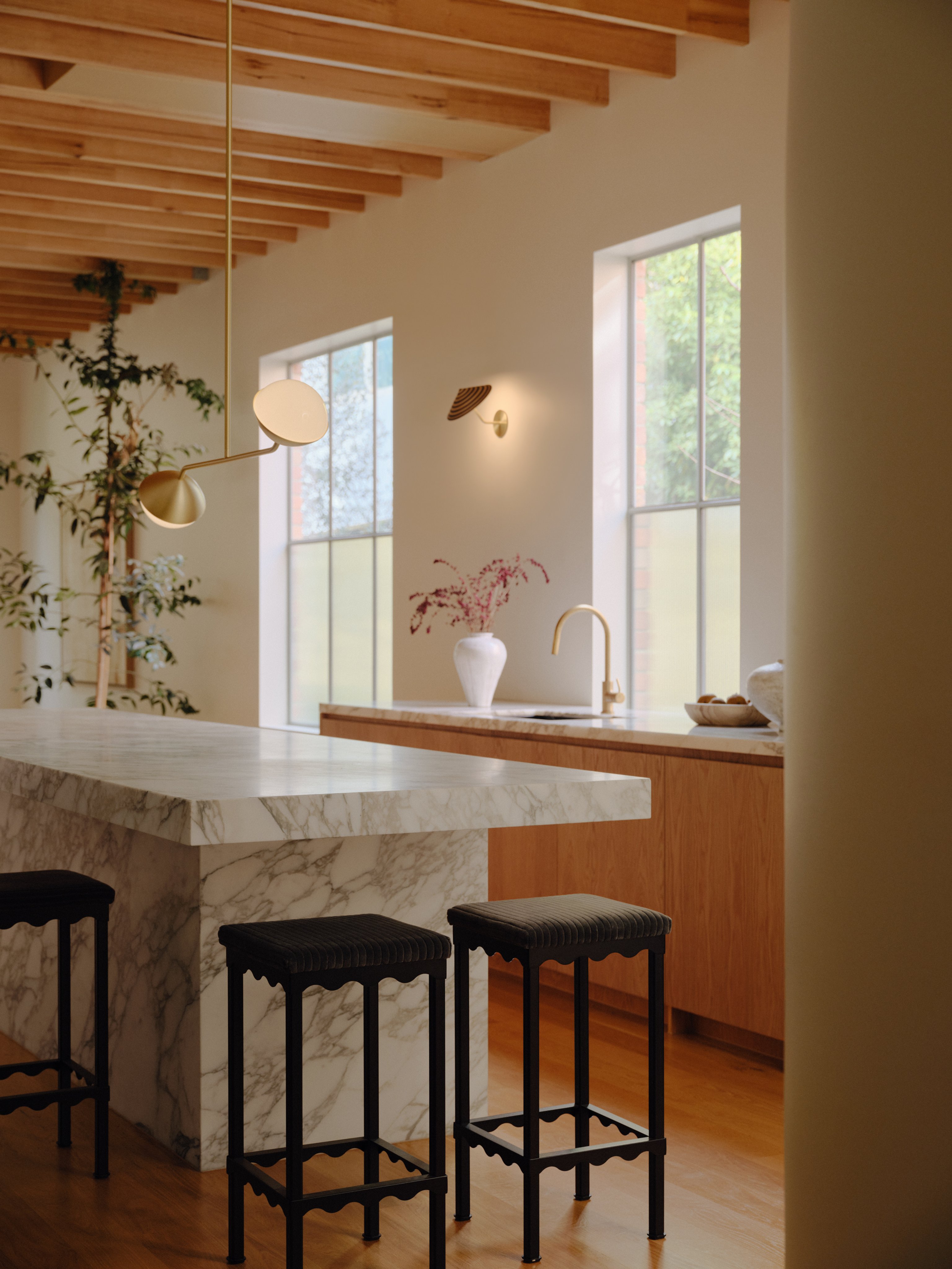 A Jolly double rod pendant in brass finish is hung from a timber ceiling in a white and timber kitchen with a marble island bench