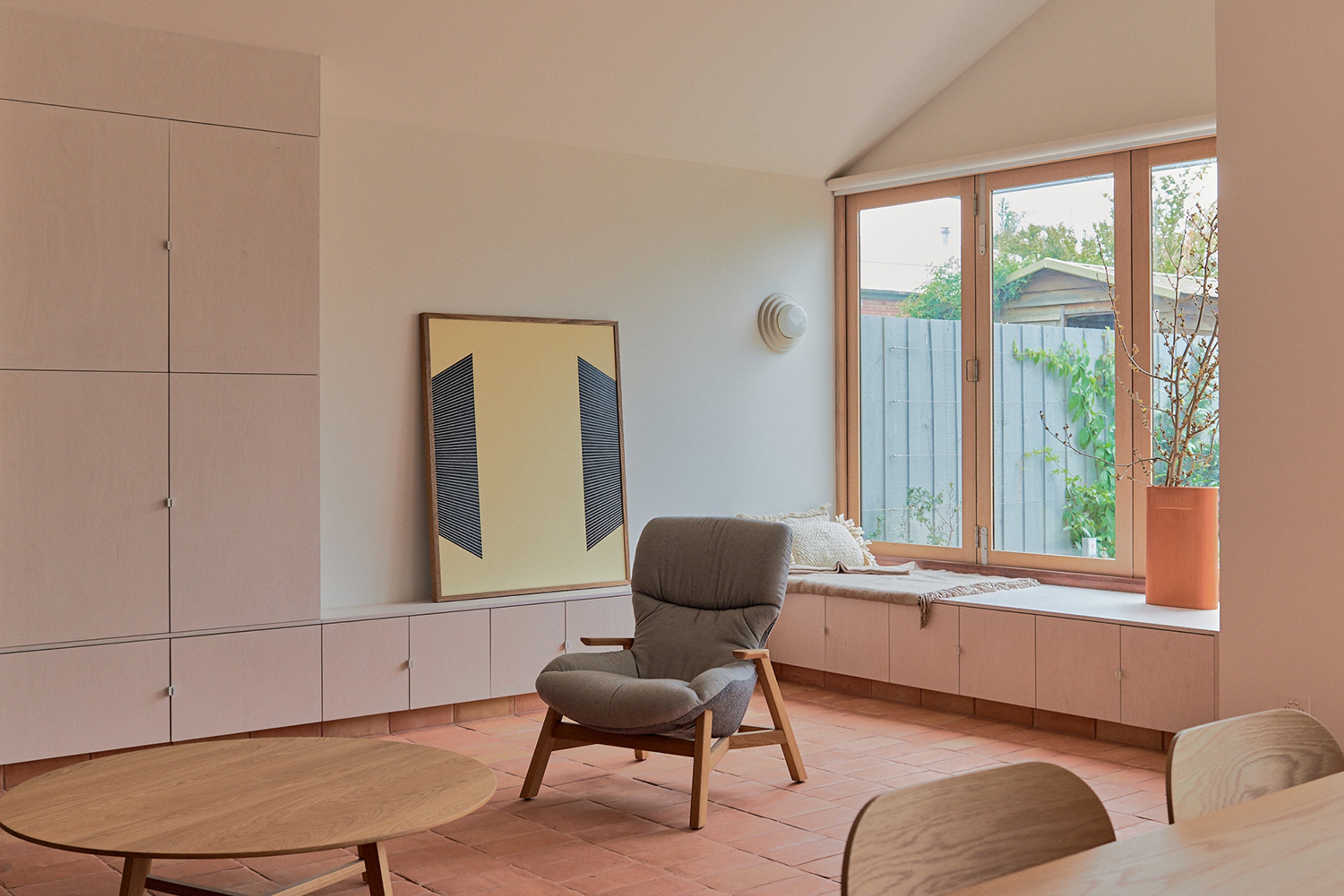 A living room with terracotta floor tiles, custom cabinetry and a large yellow artwork. Room is filled with minimalist furniture.