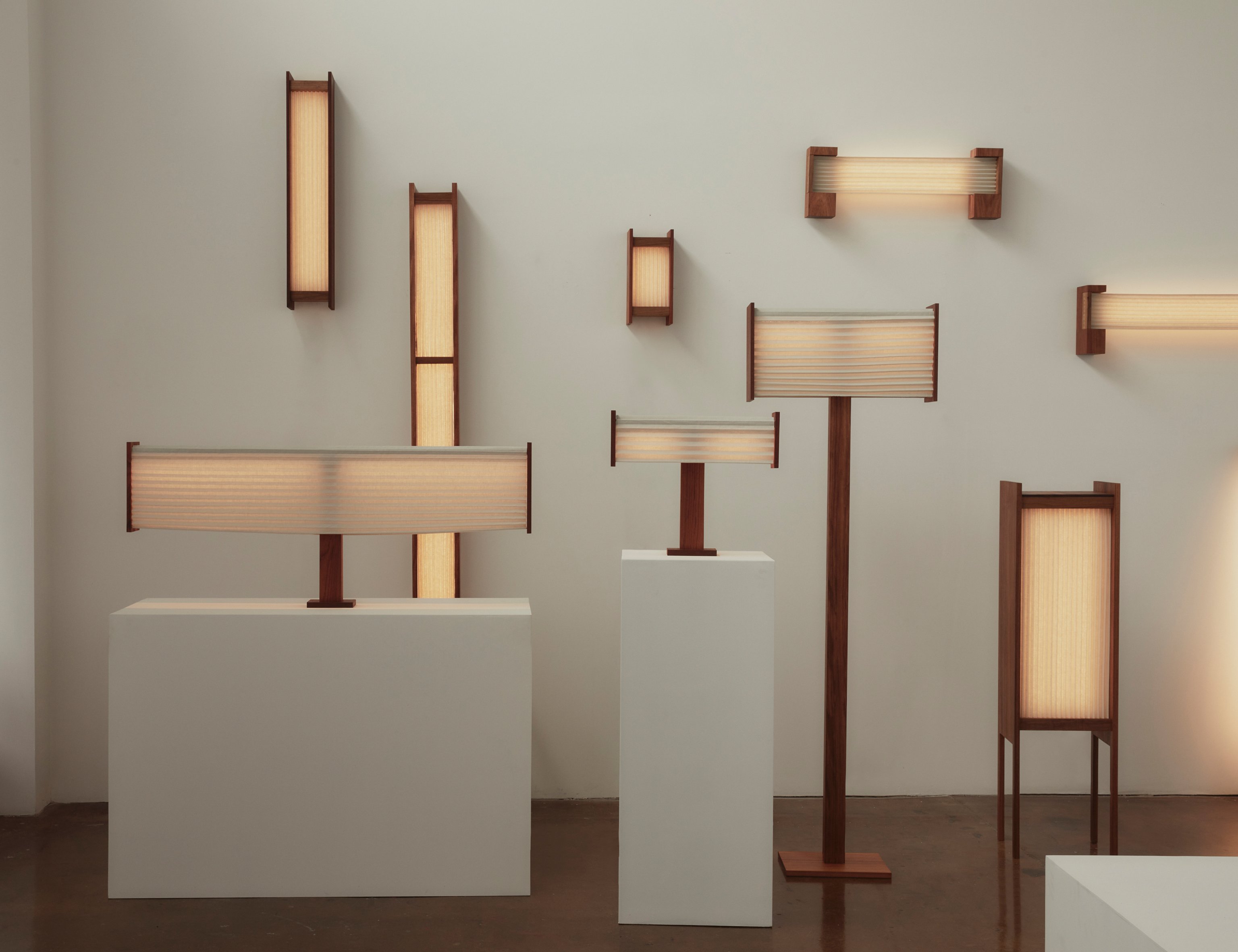 A collection of contemporary pleated linen wall light sculptures and floor lamps with timber base and warm glow, standing upright against a plain background.