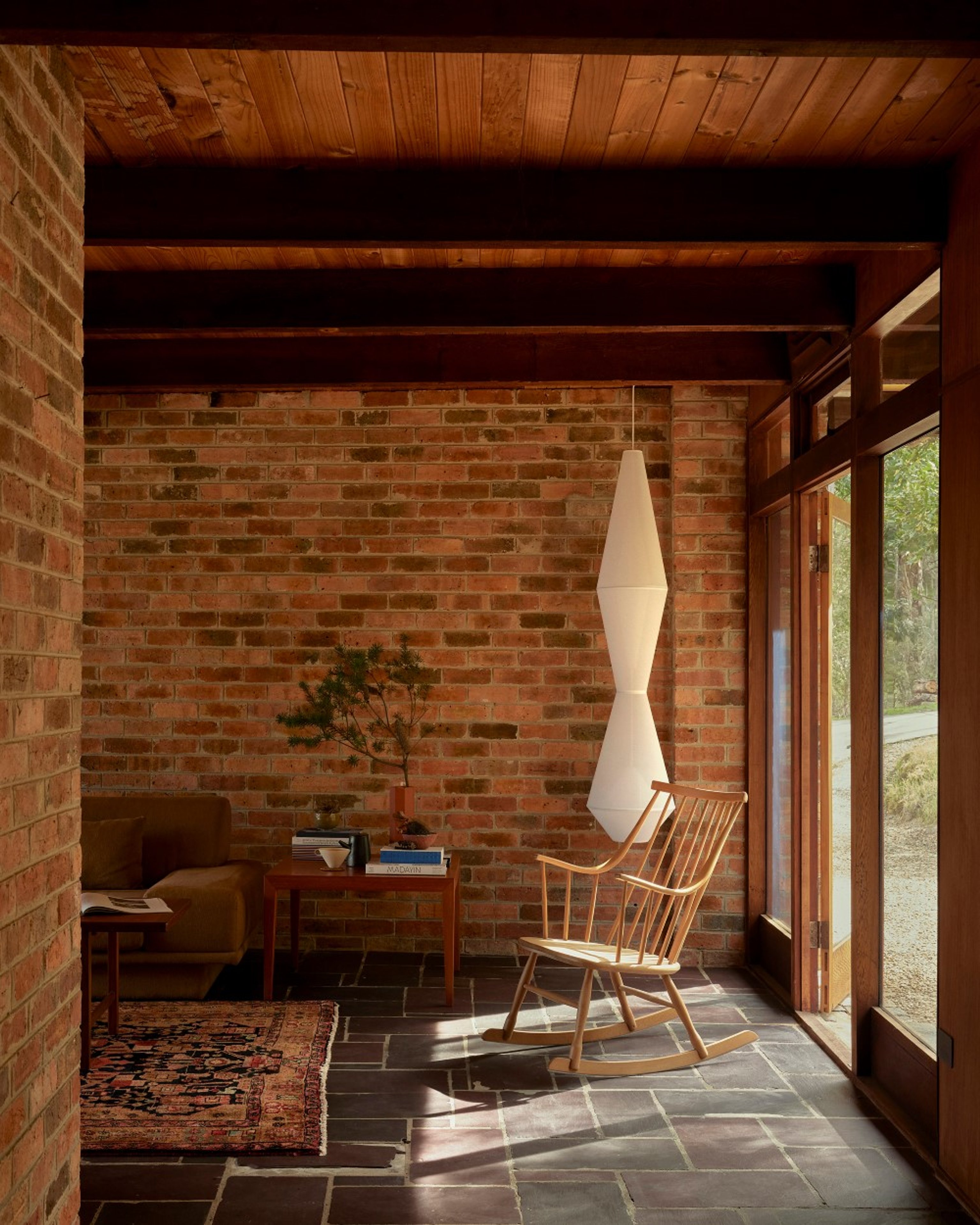 A comfortable corner of a living room with a wooden rocking chair and large fabric pendant light. The living room has stone tile floors and brick walls