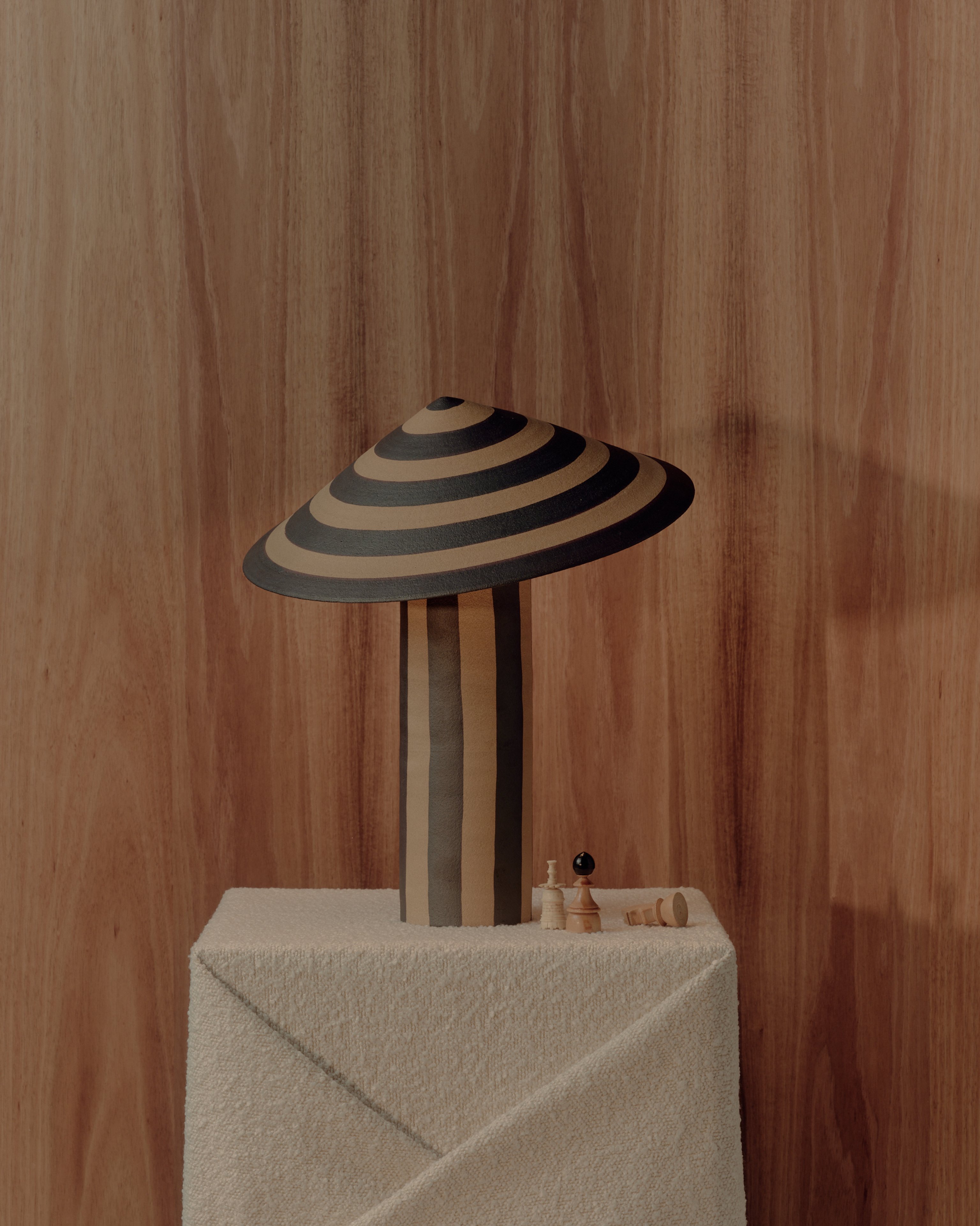 A striped ceramic table lamp sitting against a dark timber wall on a folded fabric plinth with vintage chess pieces sitting beside it.