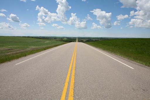 A long road through green fields, under a partly-cloudy sky