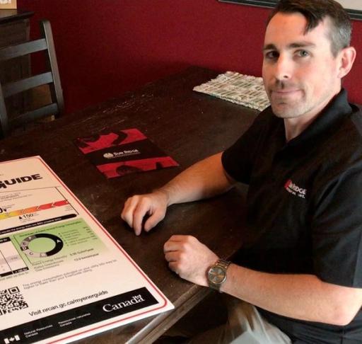 A man in a black shirt sits at a brown desk and smiles at the camera, sitting next to a variety of EnerGuide documents