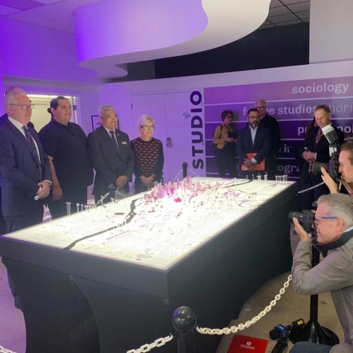 A group of four people pose in front of a city plan, while photographers and reporters stand nearby