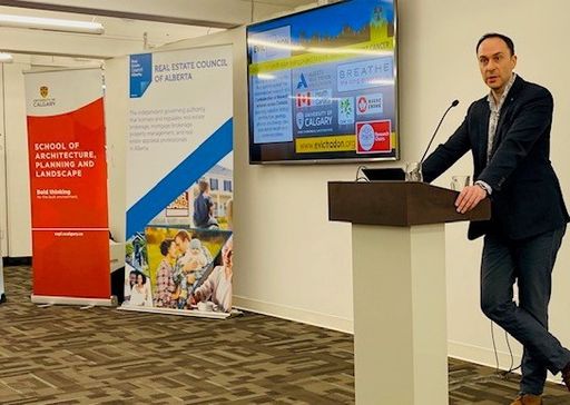 A man in a suit stands at a pedestal, presenting for the Real Estate Council of Alberta