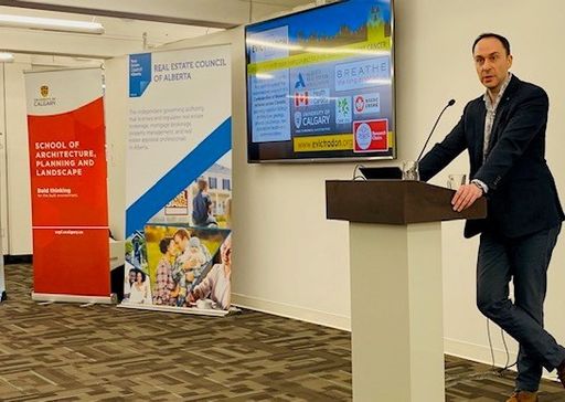 A man in a suit stands at a pedestal, presenting for the Real Estate Council of Alberta