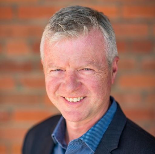 A man in a black suit jacket and blue shirt smiles at the camera, standing against a red brick wall.