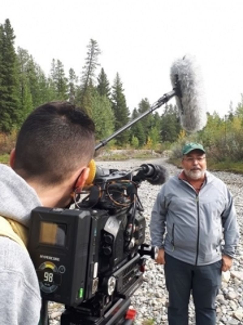 A man in a grey sweater and green hat is interviewed by someone in the forest