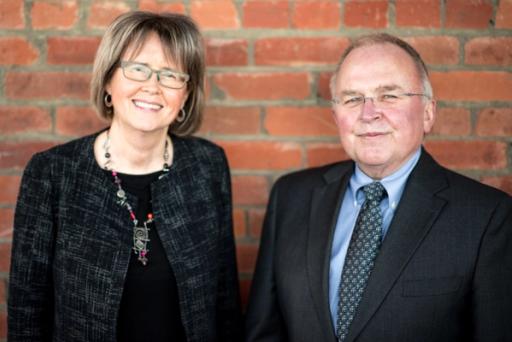 A woman wearing black and a man wearing a blue shirt and black suit jacket smile for the camera against a red brick backdrop.