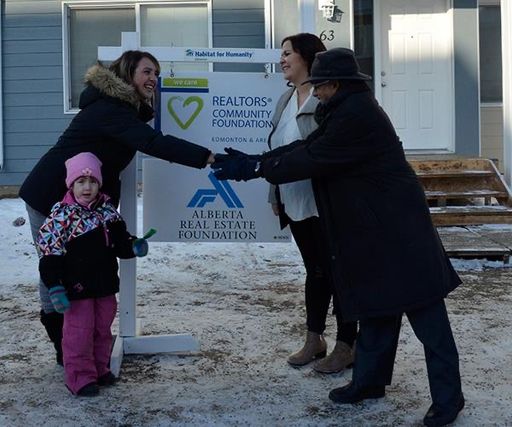 A woman with a child wearing a pink toque shakes hands with representatives from Habitat for Humanity in front of a new home.