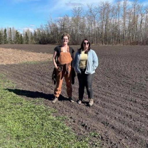 Two women stand in a dirt field with trees in the background