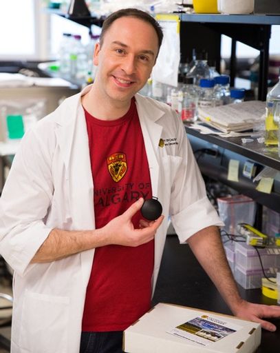 A researcher in a white lab coat and red University of Calgary shirt holds up a black ball for the camera
