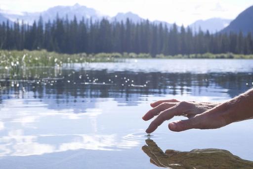 A person dips their finger into a gentle pond, with mountains and trees in the background.