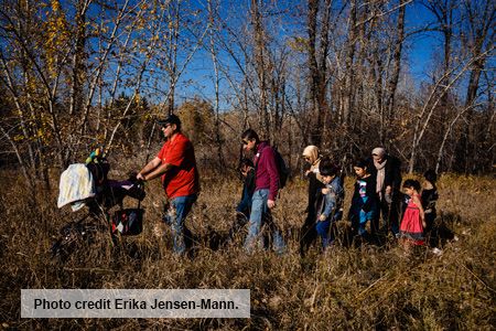A group of 8 people push a baby carriage through deep grass near a forest.