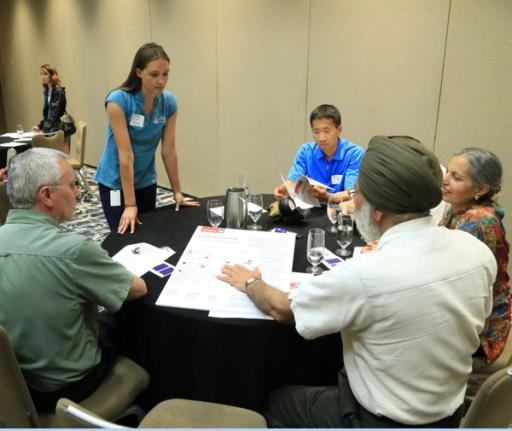 A team of five people work around a table covered in documents and coffee, while a single person stands in the background.