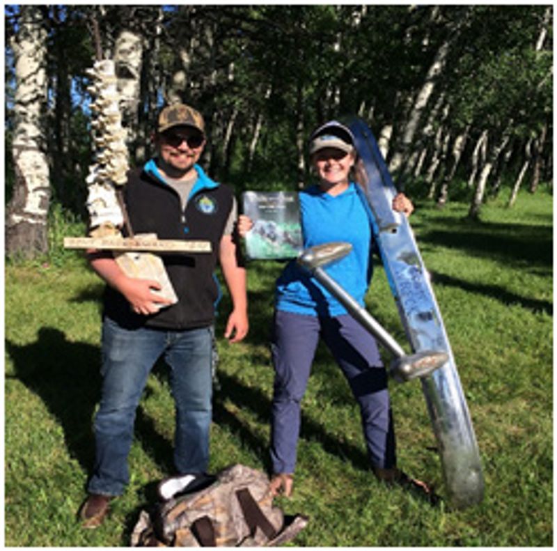 Two people holding various tools stand outside holding a document and smiling at the camera