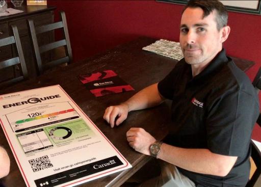 A man in a black shirt sits next to a large 'EnerGuide' sign