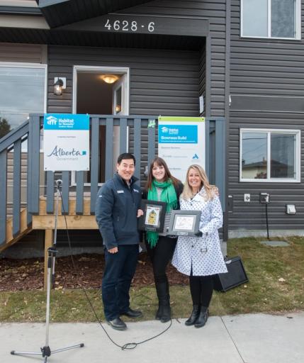 Three people stand in front of a house with black siding, holding up plaques for Habitat for Humanity.