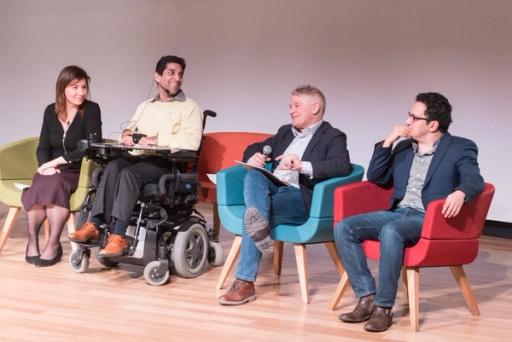 A panel of four people, three men and one woman, one in a wheelchair, discuss a topic on a wooden stage.