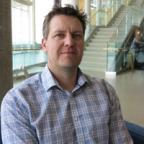 A man in a plaid shirt stairs in a contemplative manner directly at the camera, in front of a set of stairs