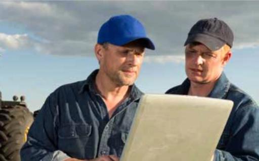 Two farmers wearing baseball caps review details on a durable sand-colored laptop