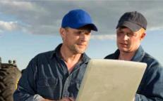 Two farmers wearing baseball caps review details on a durable sand-colored laptop