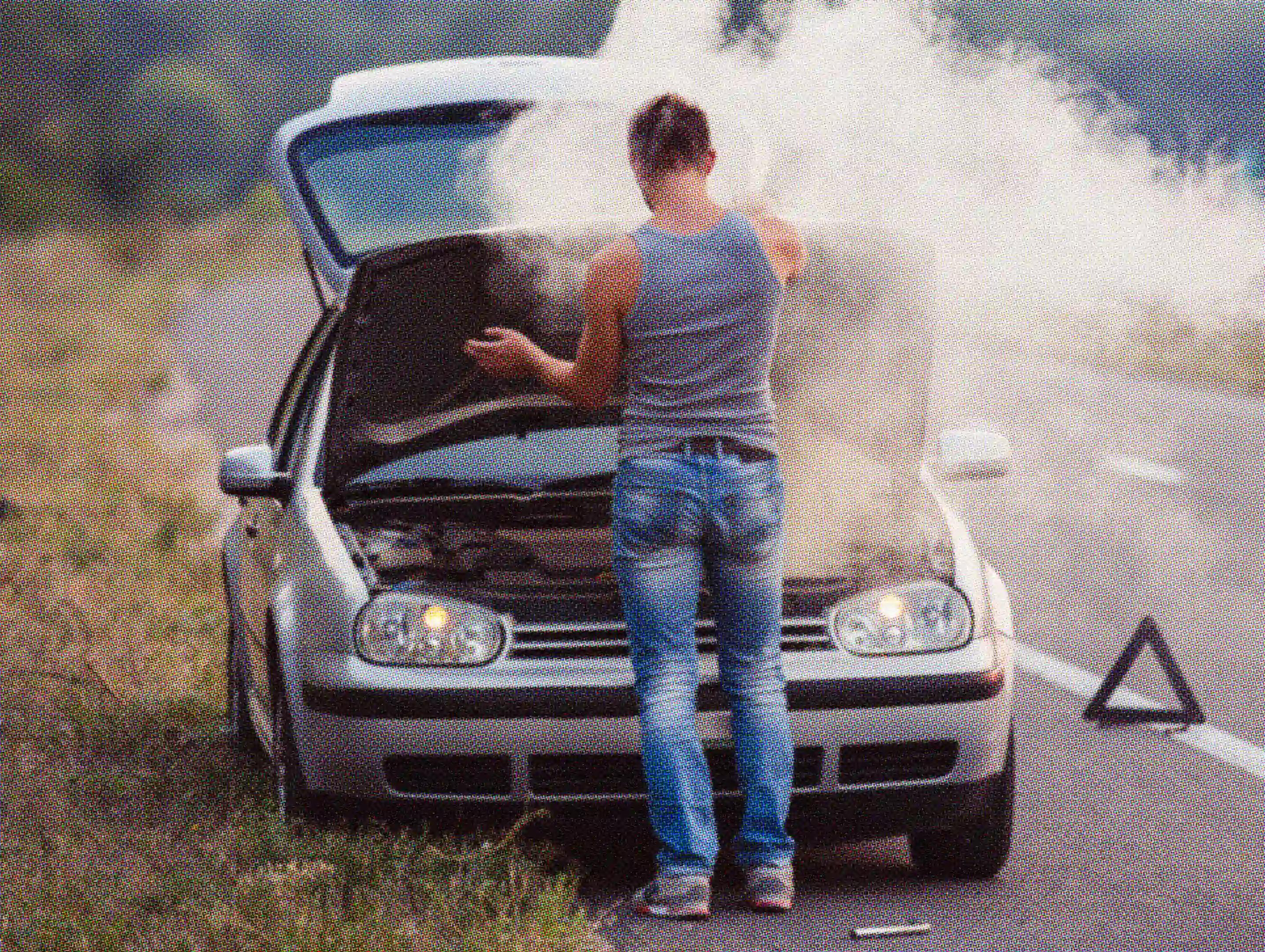 A person standing in front of a smoking car with the hood up on the side of a road, signaling a breakdown.