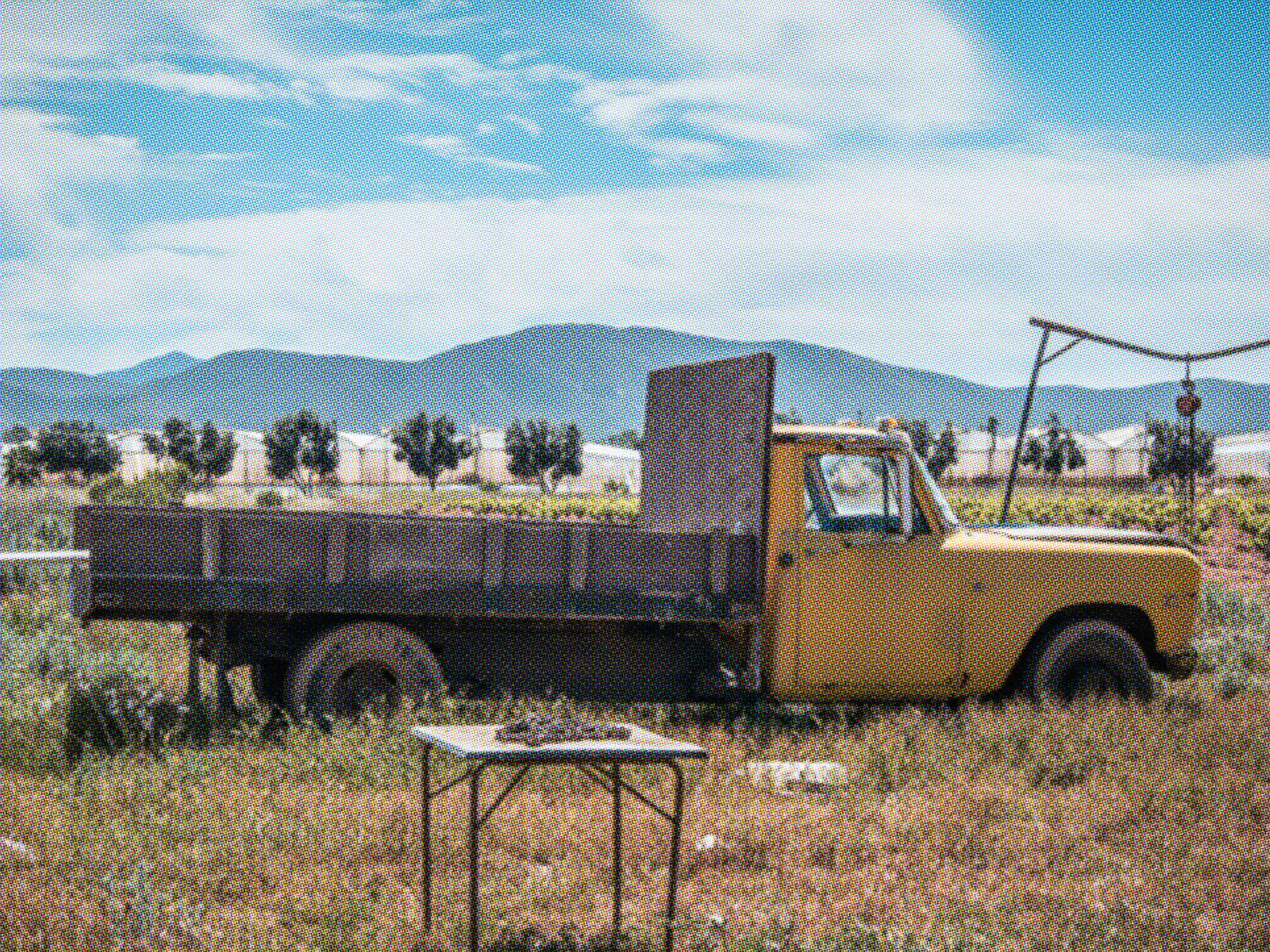 Vintage yellow flatbed work truck parked in a dry desert field with mountains in the background, showing signs of age and wear.