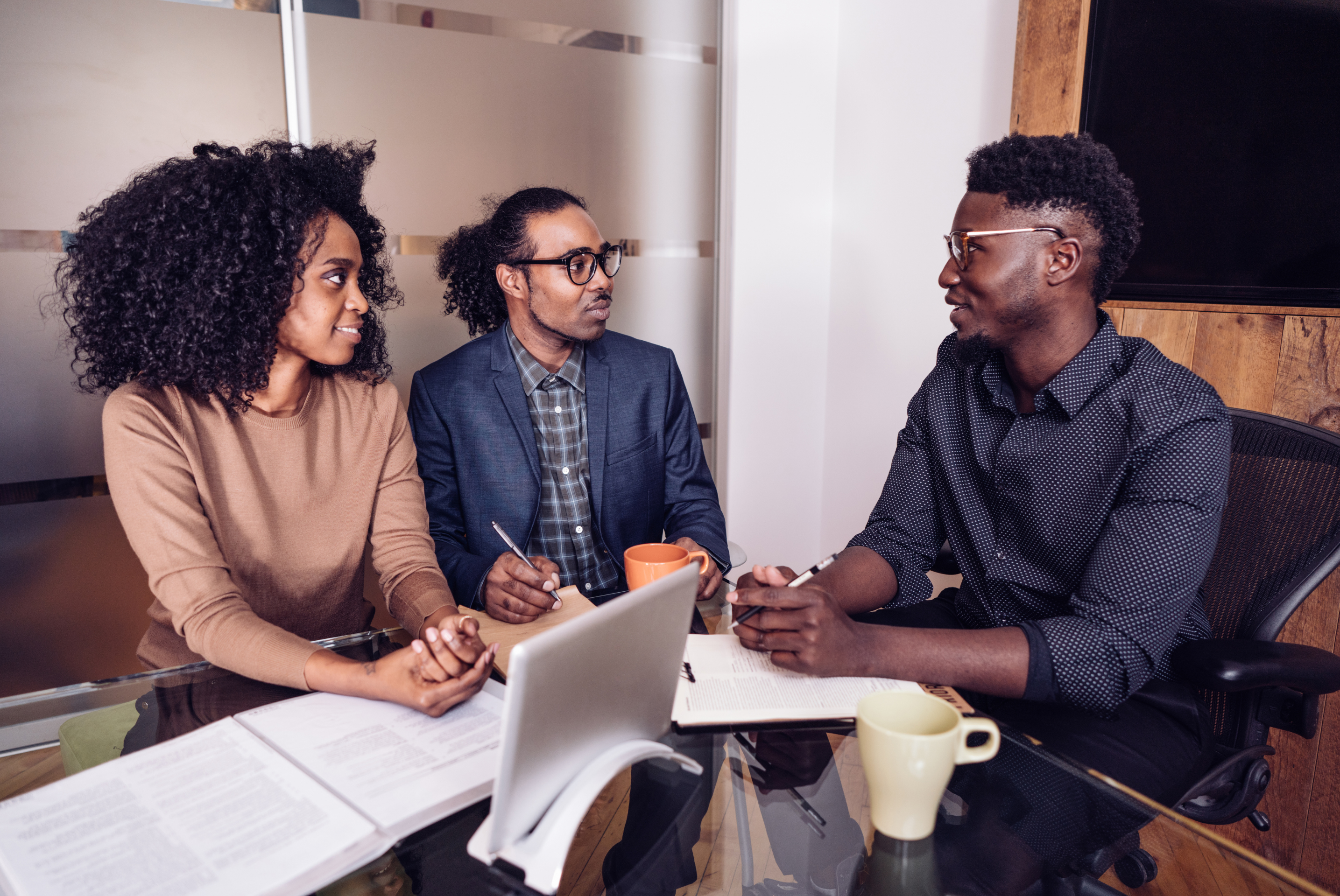 "Three professionals discussing paperwork during a funding meeting, taking notes around a glass table